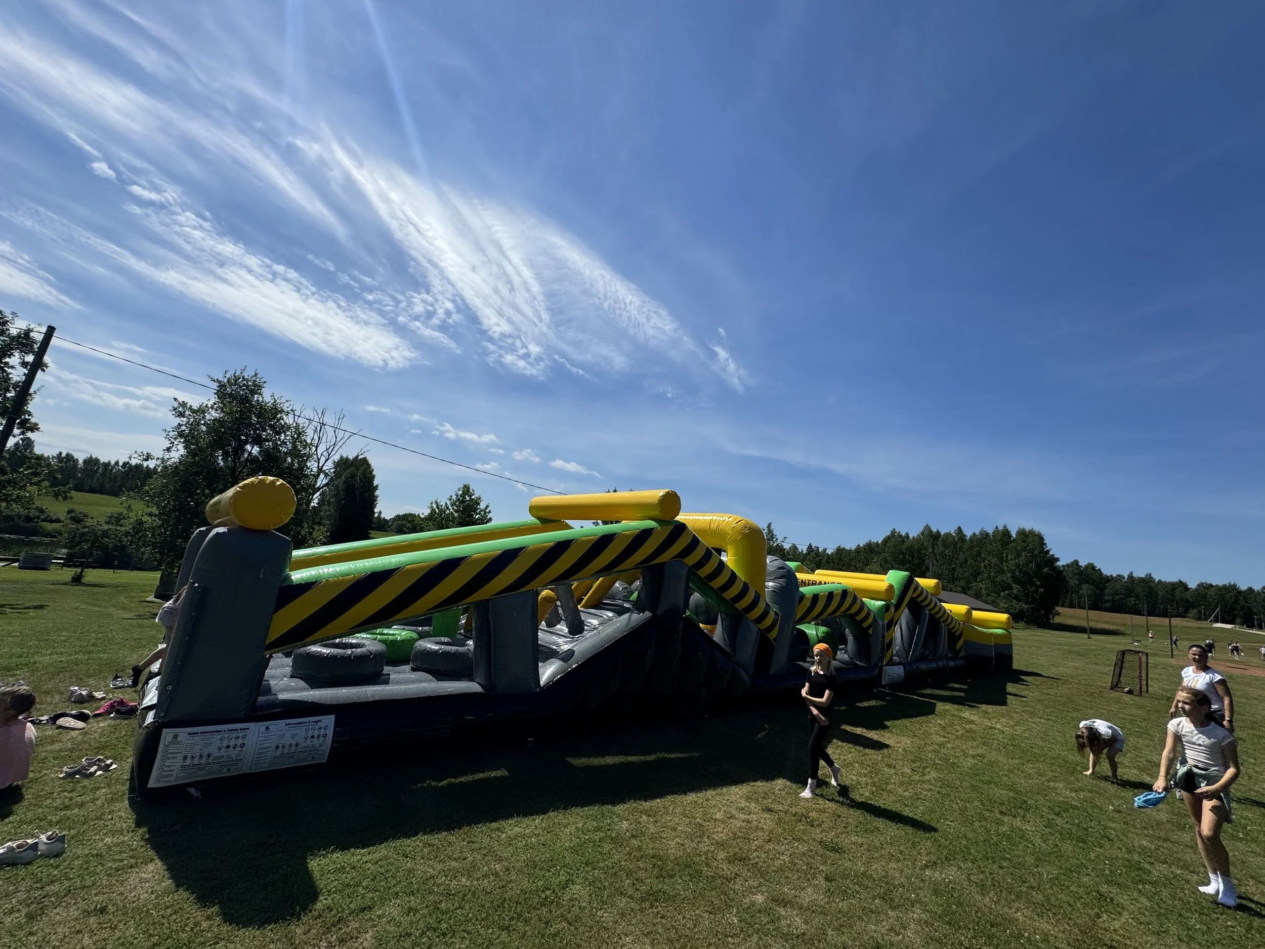 Large inflatable obstacle course set up outdoors on grass under a blue sky with a few clouds, with children and adults nearby.