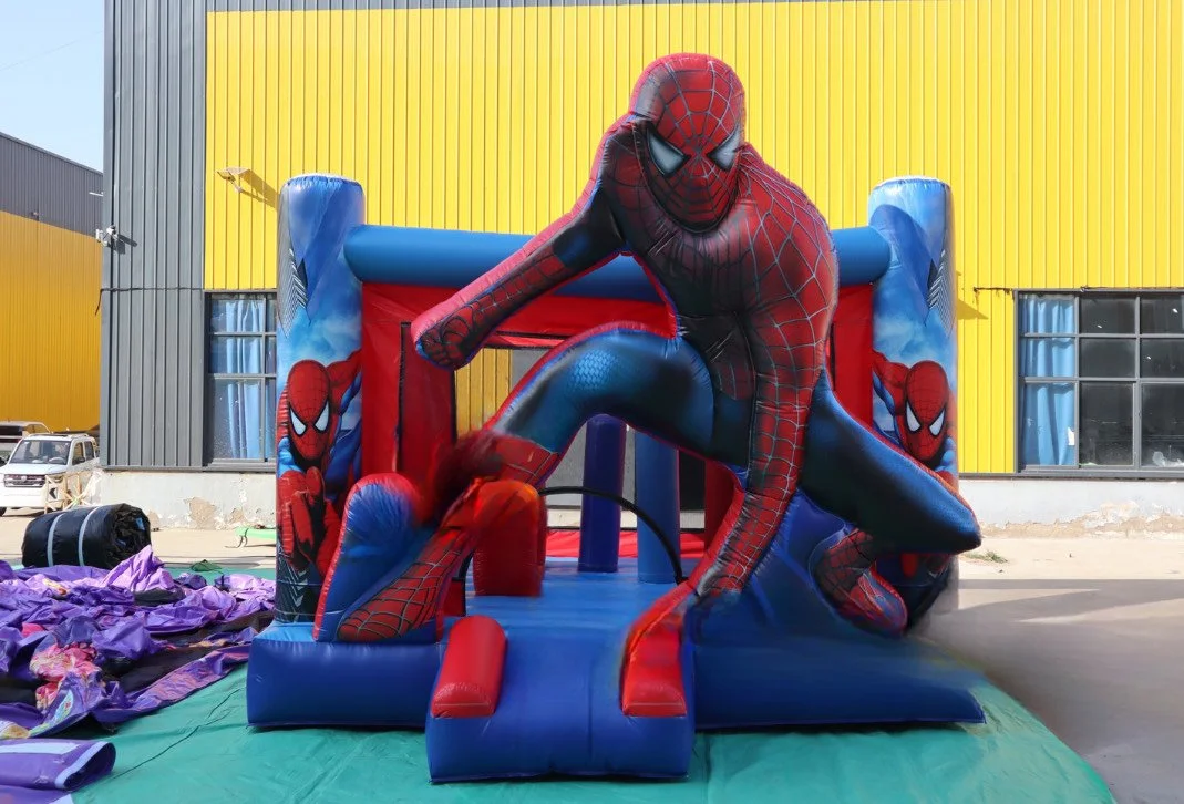 A Spider-Man themed inflatable bounce house set outside, with a large Spider-Man figure climbing on it, positioned in front of a yellow and gray building.