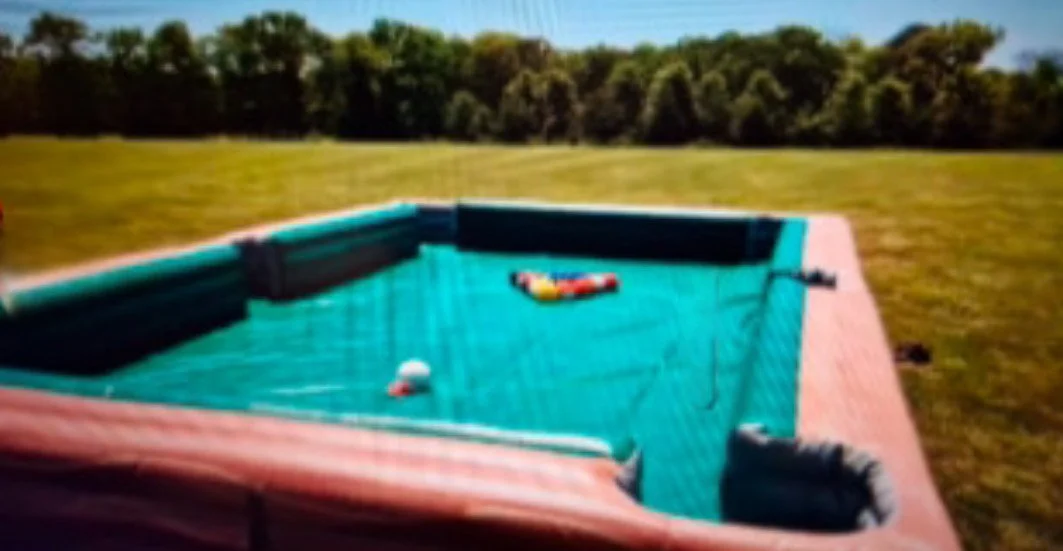 Inflatable pool filled with pool balls, set up outdoors on grass with a background of trees and blue sky.