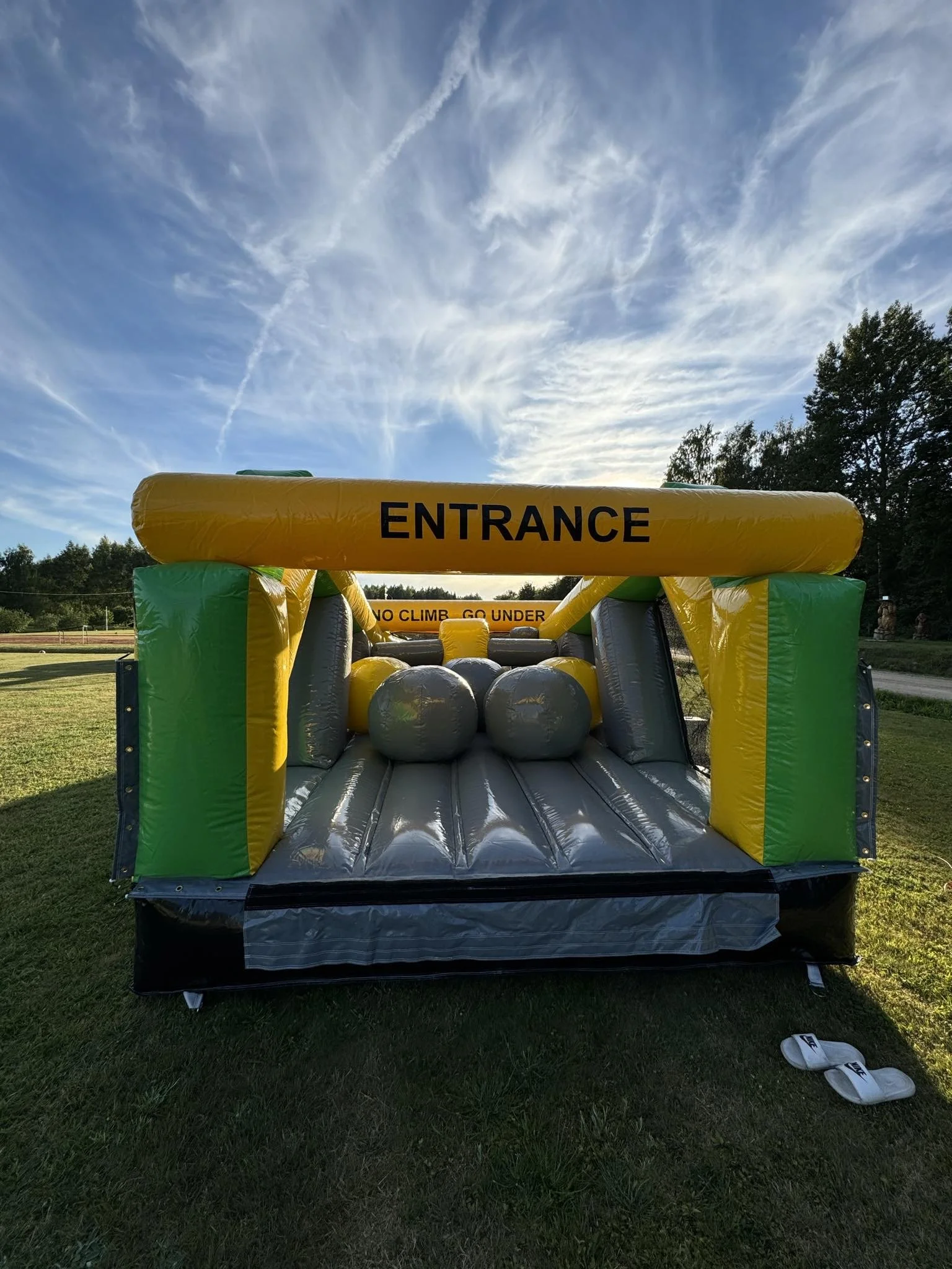Colorful inflatable bounce house labeled 'ENTRANCE' with a warning sign 'NO CLIMB GO UNDER' inside, set up outdoors on grass with trees in the background and a partly cloudy sky overhead.