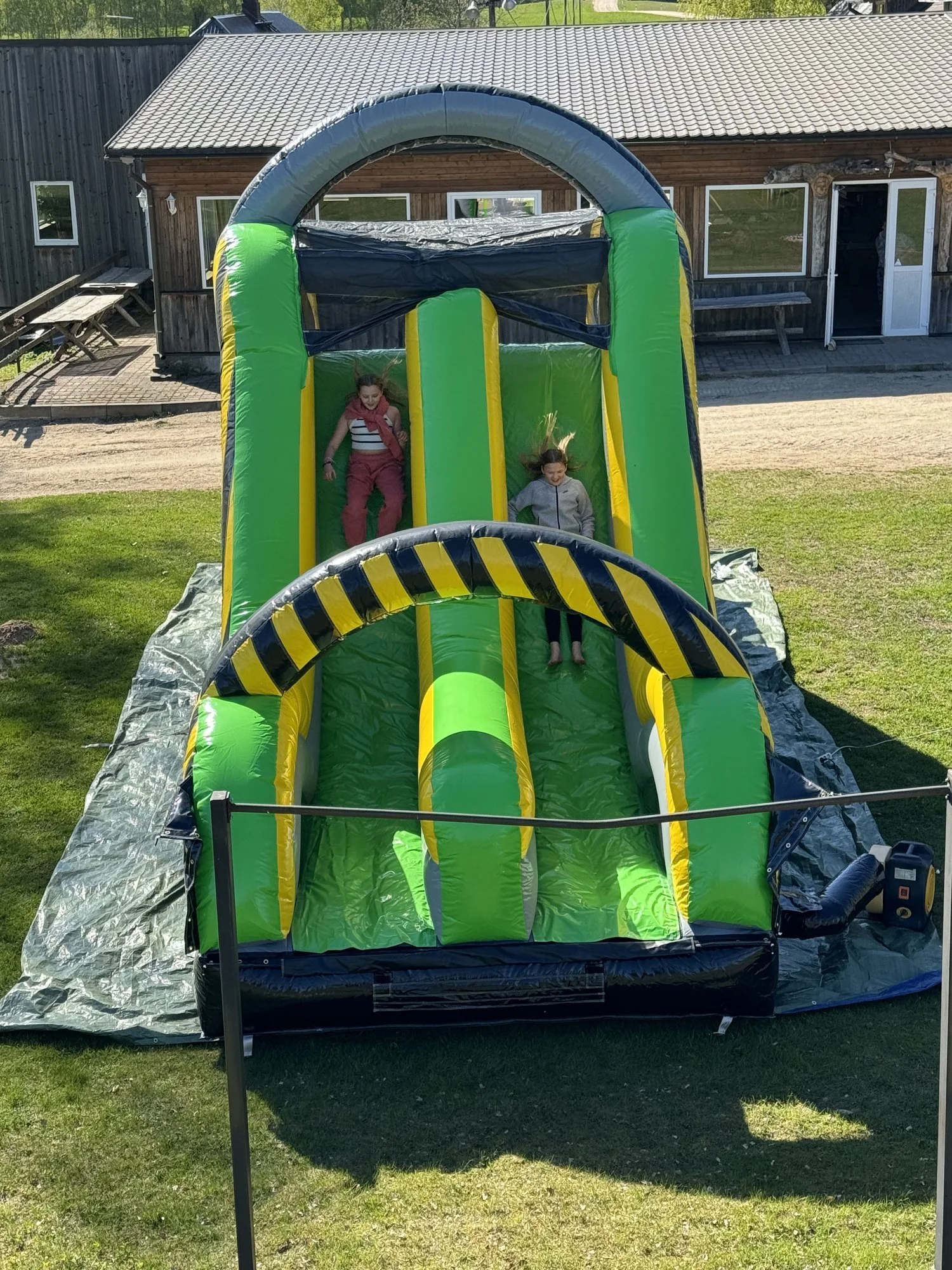 Children playing on a large inflatable slide with green, yellow, and black colors, set up on grass outside a wooden building.