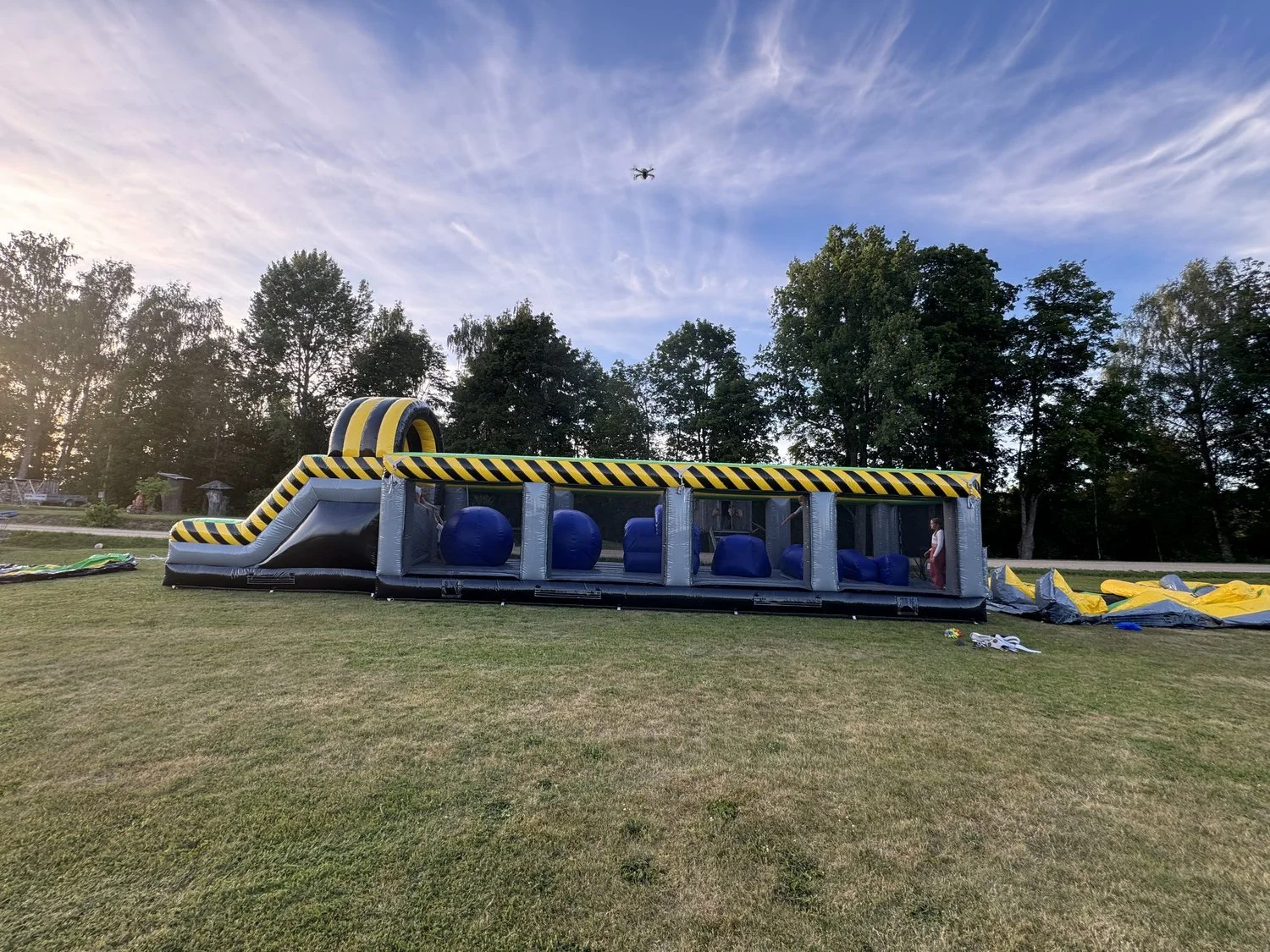 An indoor inflatable obstacle course set up outdoors on a grassy field with trees in the background and a drone flying overhead.