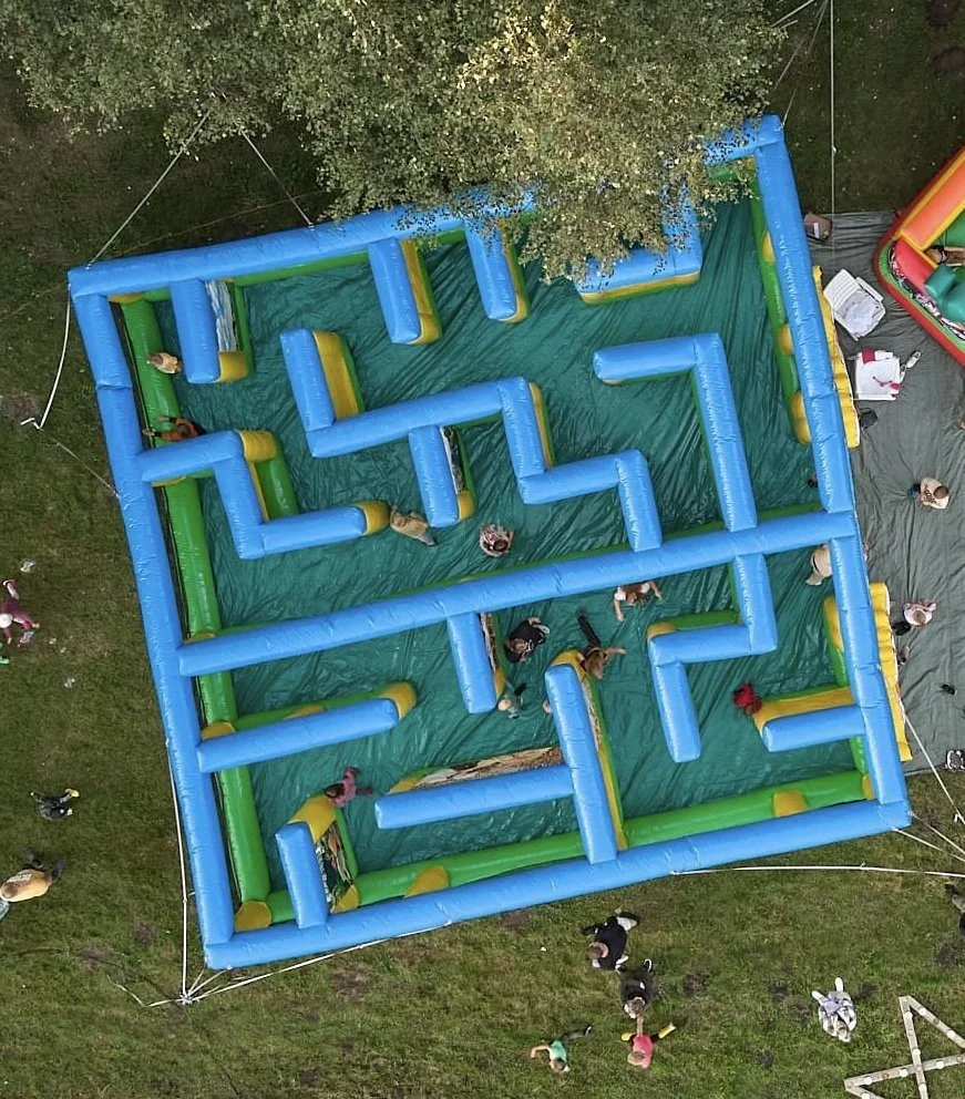 Overhead view of a large inflatable maze with blue and green walls on a grassy area, with children playing inside and adults watching outside.