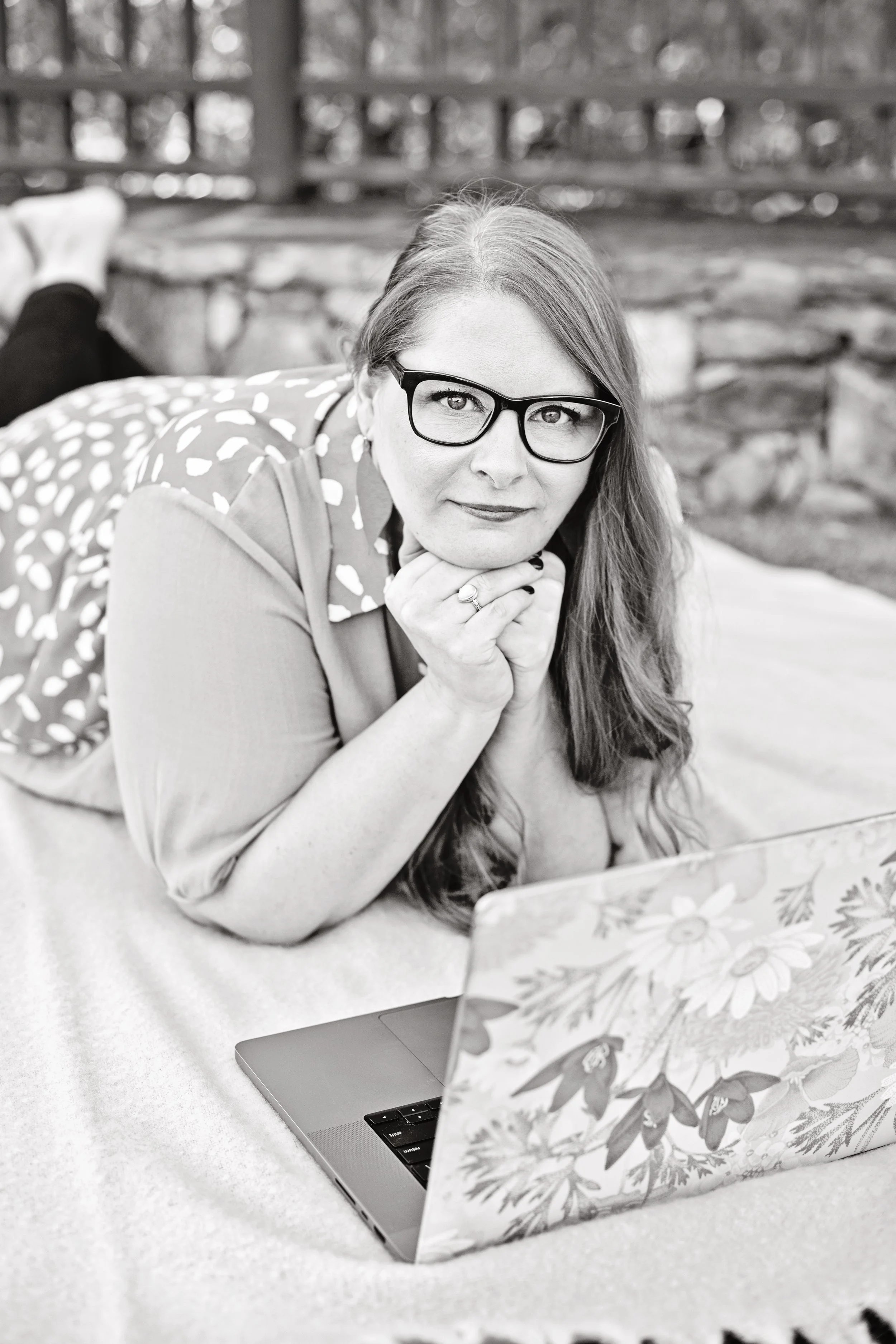 A woman with glasses lying on her stomach on a couch, resting her chin on her hands, with a laptop decorated with a floral pattern in front of her, outdoors with a stone wall and wooden fence in the background.