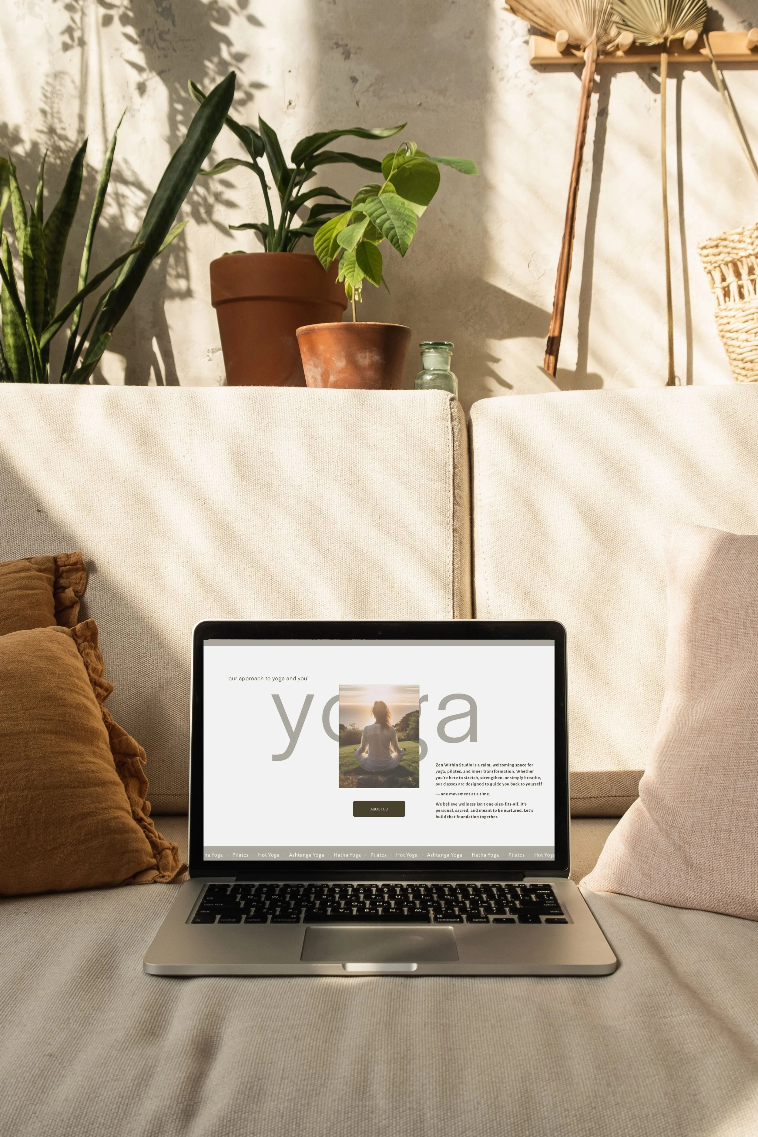 Laptop on beige couch with pillows, showing a yoga website, in a cozy room with potted plants and natural sunlight.