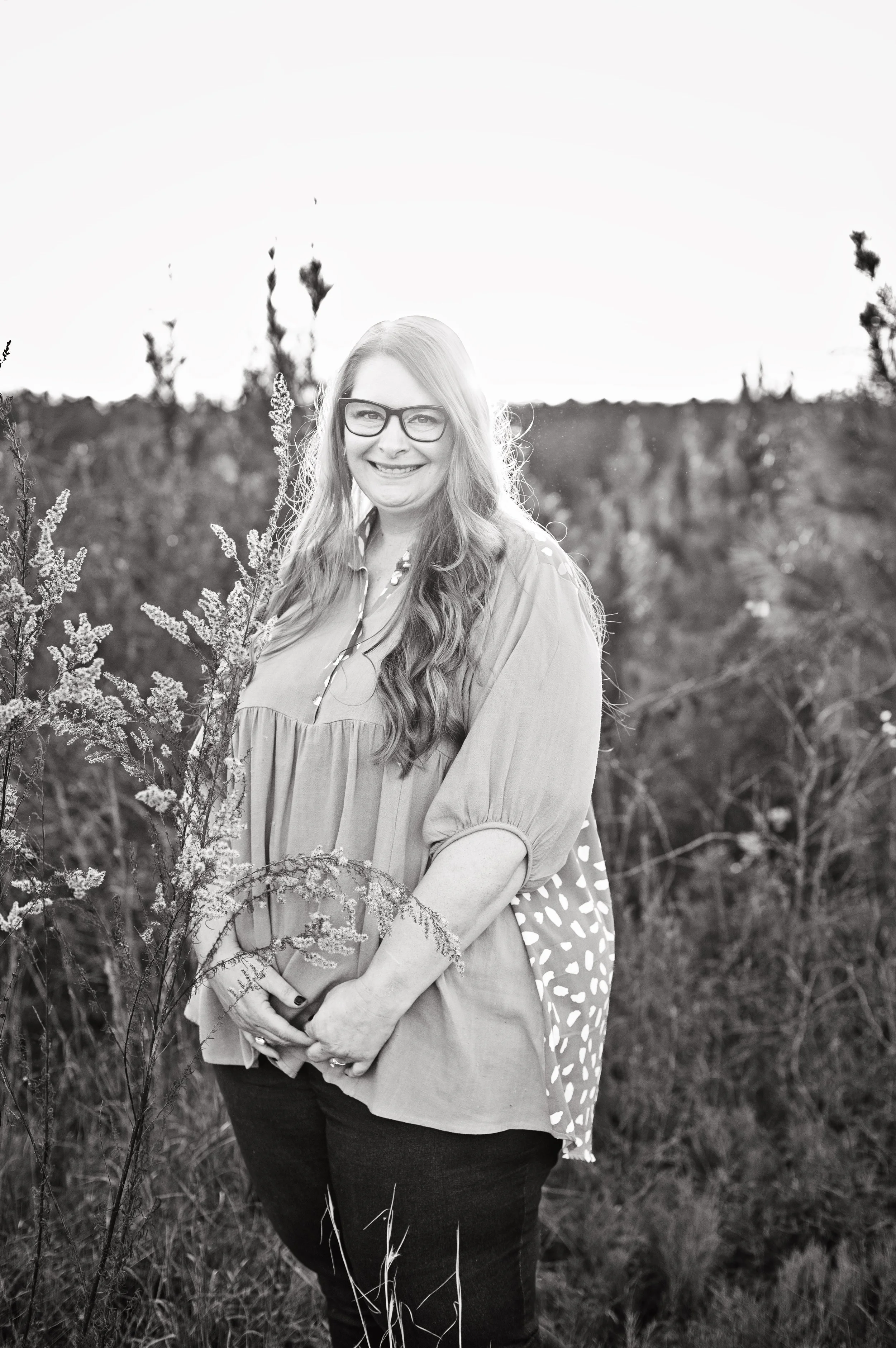 A smiling woman with long wavy hair and glasses standing outside in a field with tall grass and plants, wearing a loose blouse and dark pants.