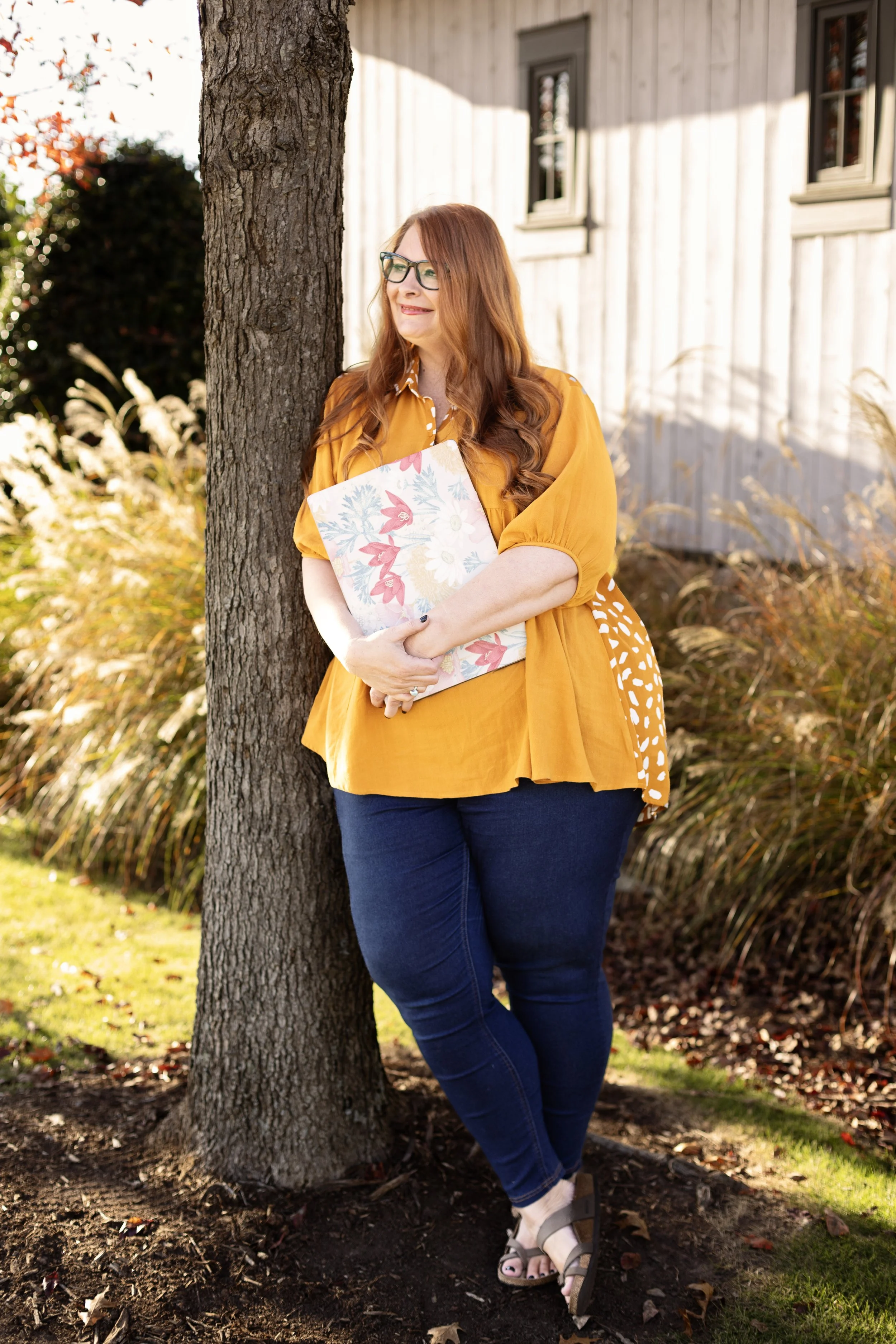 A woman with red hair and glasses leaning against a tree in a garden, holding a floral notebook, wearing an orange top and dark jeans.
