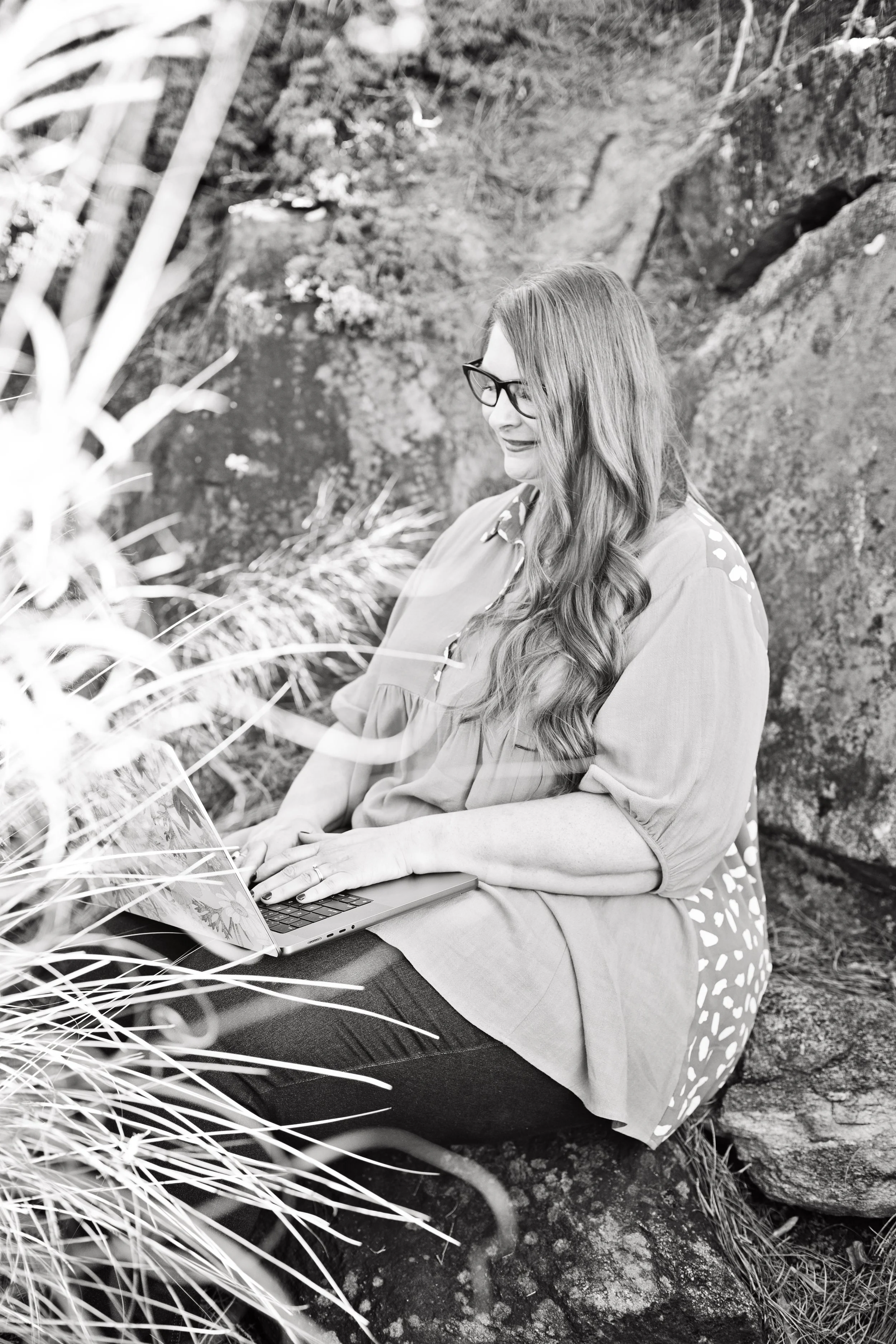 Black and white photo of a woman with long wavy hair and glasses sitting outdoors on a rock with a laptop, surrounded by nature.