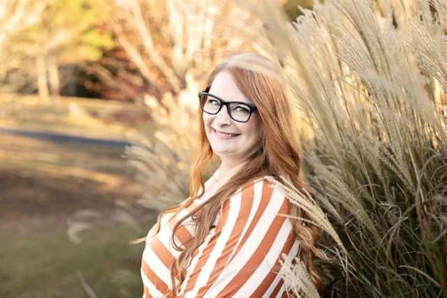 A smiling woman with long red hair and glasses standing near tall ornamental grasses outdoors during fall.