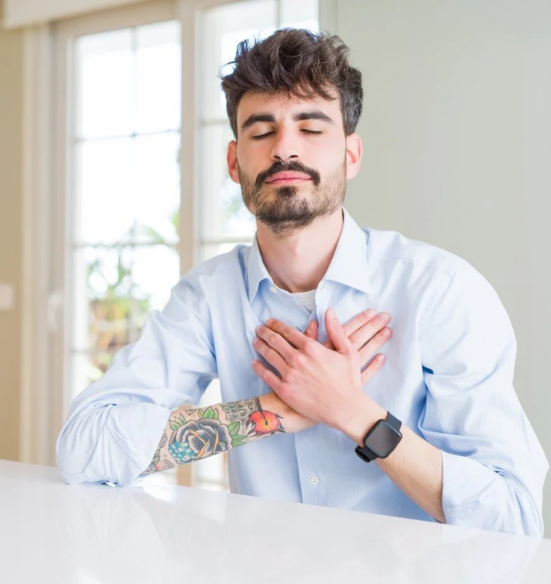 Man with closed eyes and hands on chest, wearing a blue shirt, with tattoos on one arm.