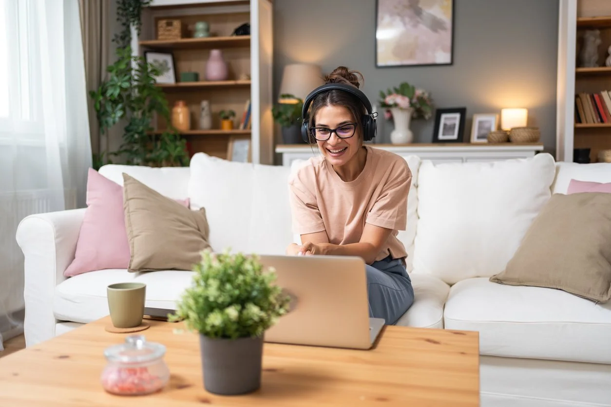 A woman sitting on a white couch in a living room, wearing headphones and smiling while looking at a laptop on a wooden coffee table. The room has decorative pillows, plants, and bookshelves in the background.
