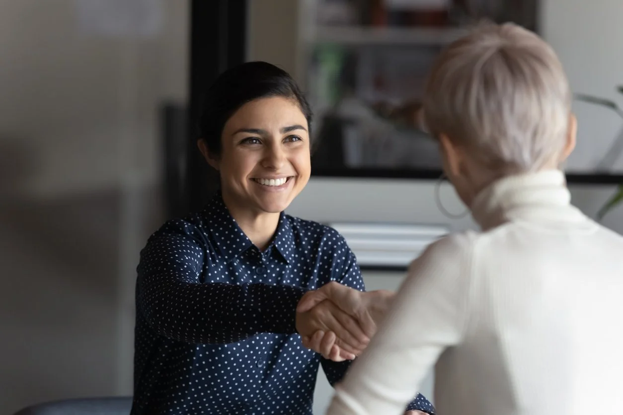Two women shaking hands in an office, one smiling and wearing a polka dot shirt, the other with short blonde hair facing away.