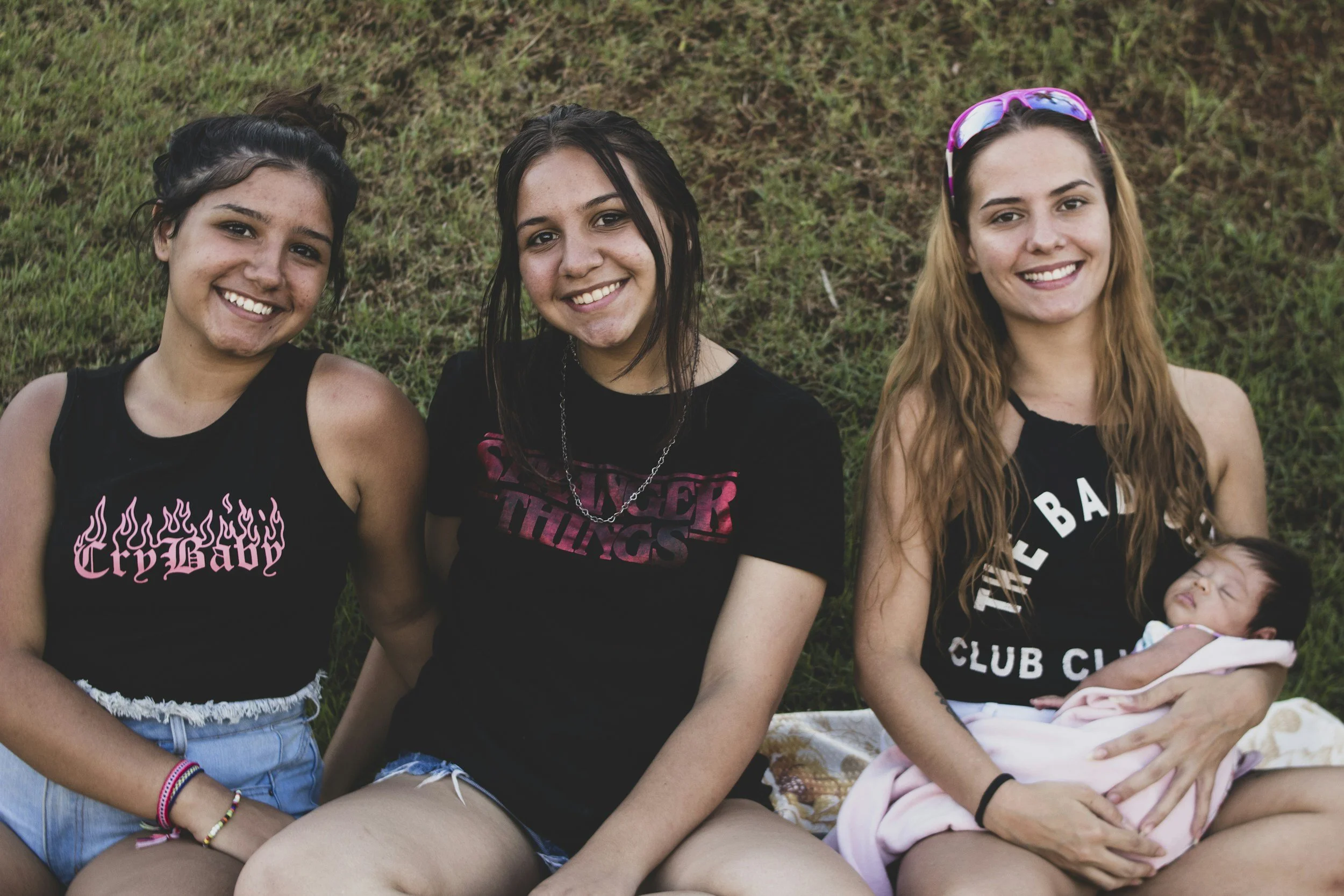 A woman with curly hair holding a baby and kissing the baby's cheek, both in white tops against a plain background.