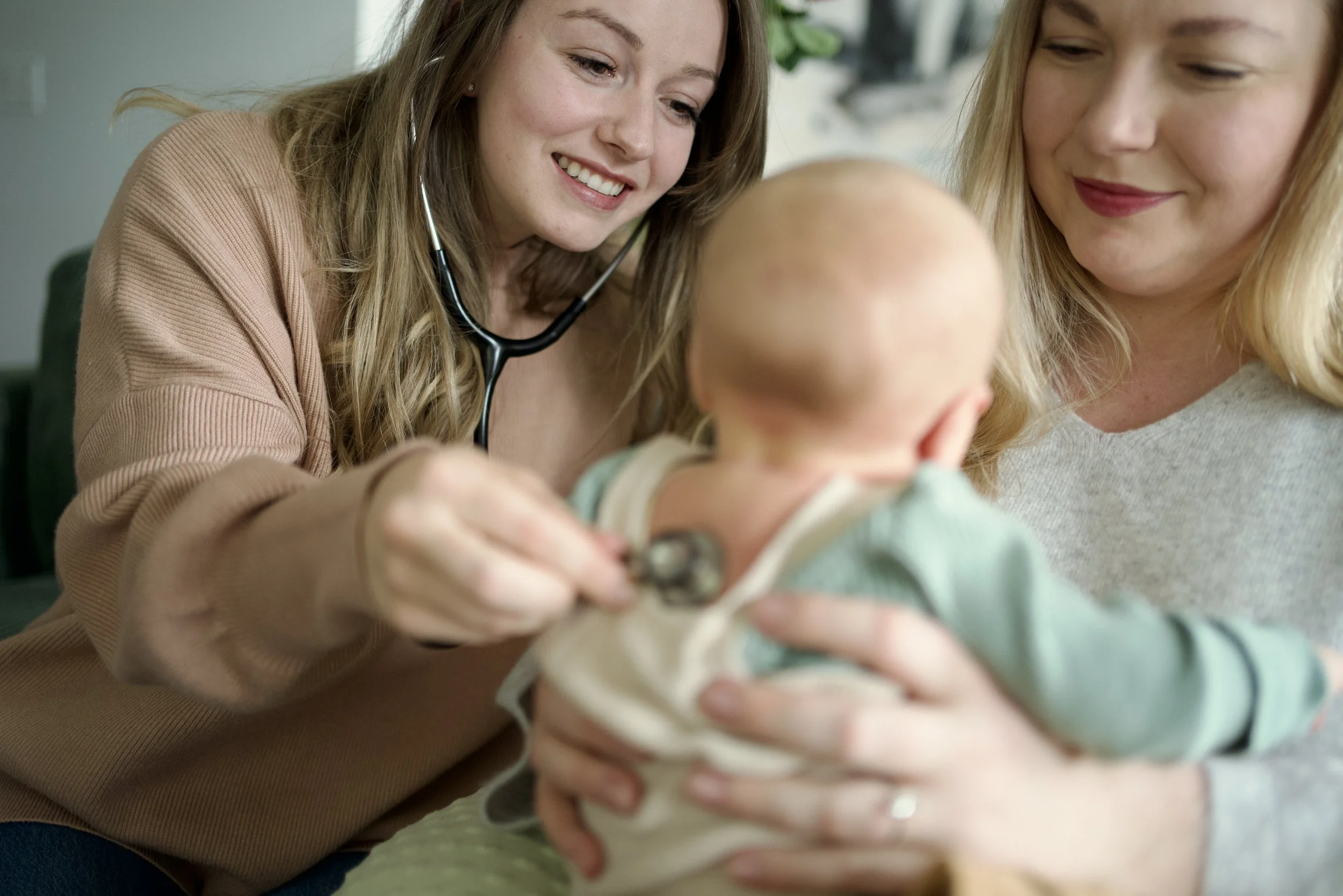 A woman breastfeeding a baby while a nurse prepares to give the baby a medical checkup, with a young girl standing beside them in a living room decorated for Christmas.