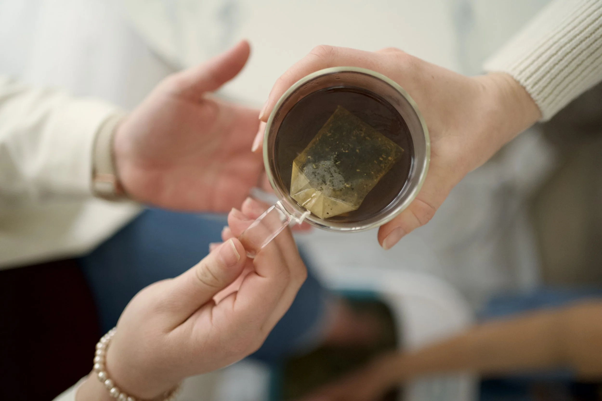 A wicker chair with a white decorative pillow, accompanied by large green indoor plants and sheer curtains in the background.