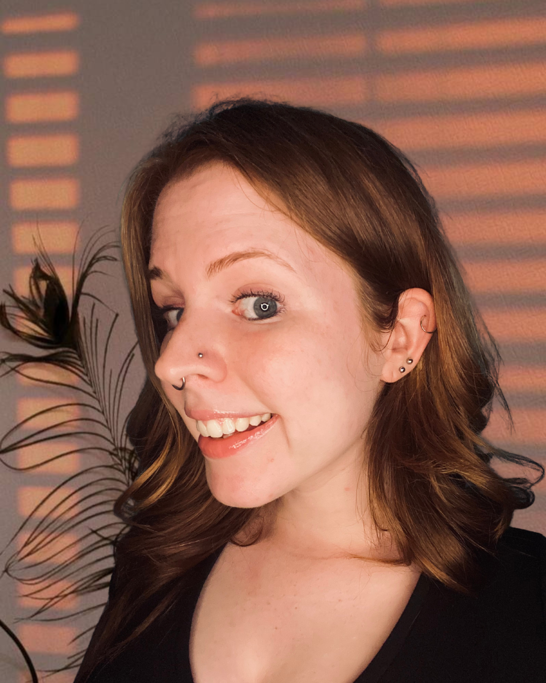 A woman with reddish-brown hair and blue eyes smiling, with a plant and sunset light shadows on the wall behind her.