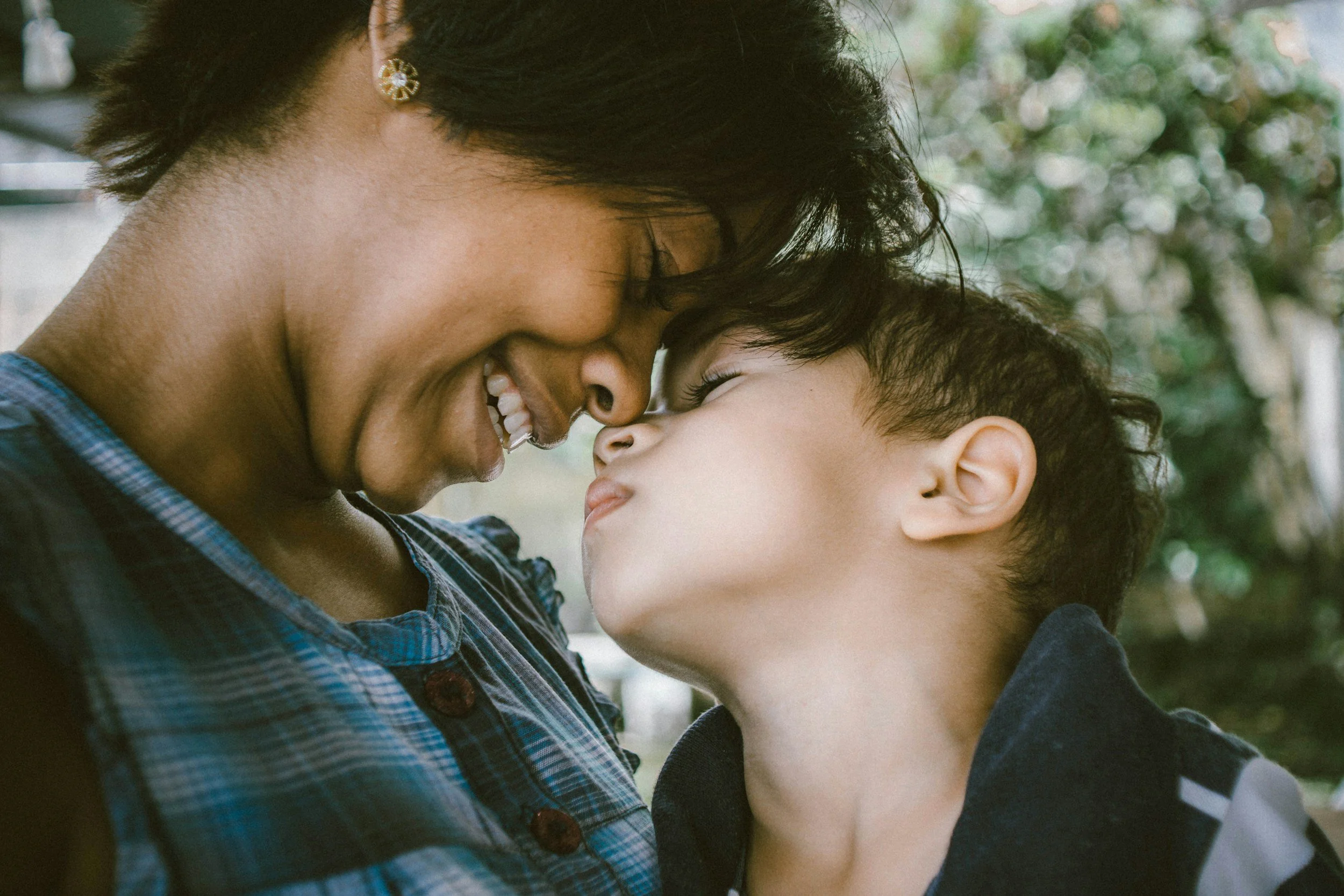 A woman and a young boy gently touching foreheads and smiling outdoors.