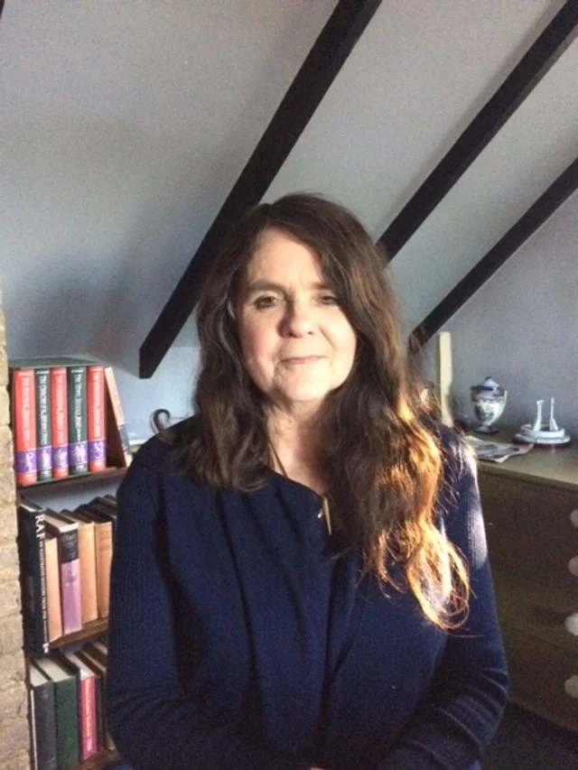 A woman with long brown hair and a dark top sitting indoors with a bookshelf and decorative items in the background under a sloped ceiling.