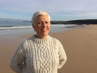 An older woman with short white hair standing on a beach, smiling, wearing a cream-colored knit sweater, with the ocean and a cloudy sky in the background.