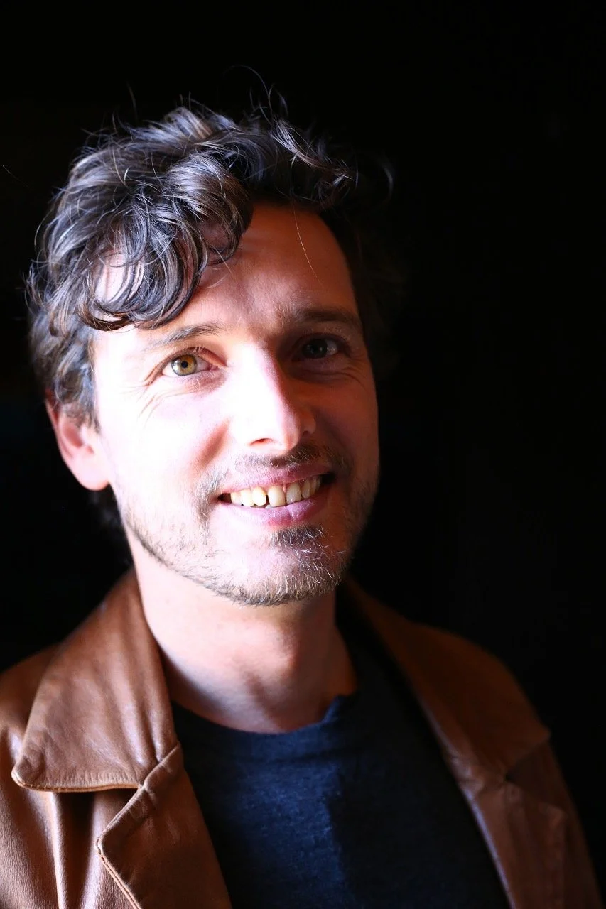 Portrait of a smiling man with wavy brown hair, light facial hair, wearing a brown leather jacket and dark shirt, against a black background.