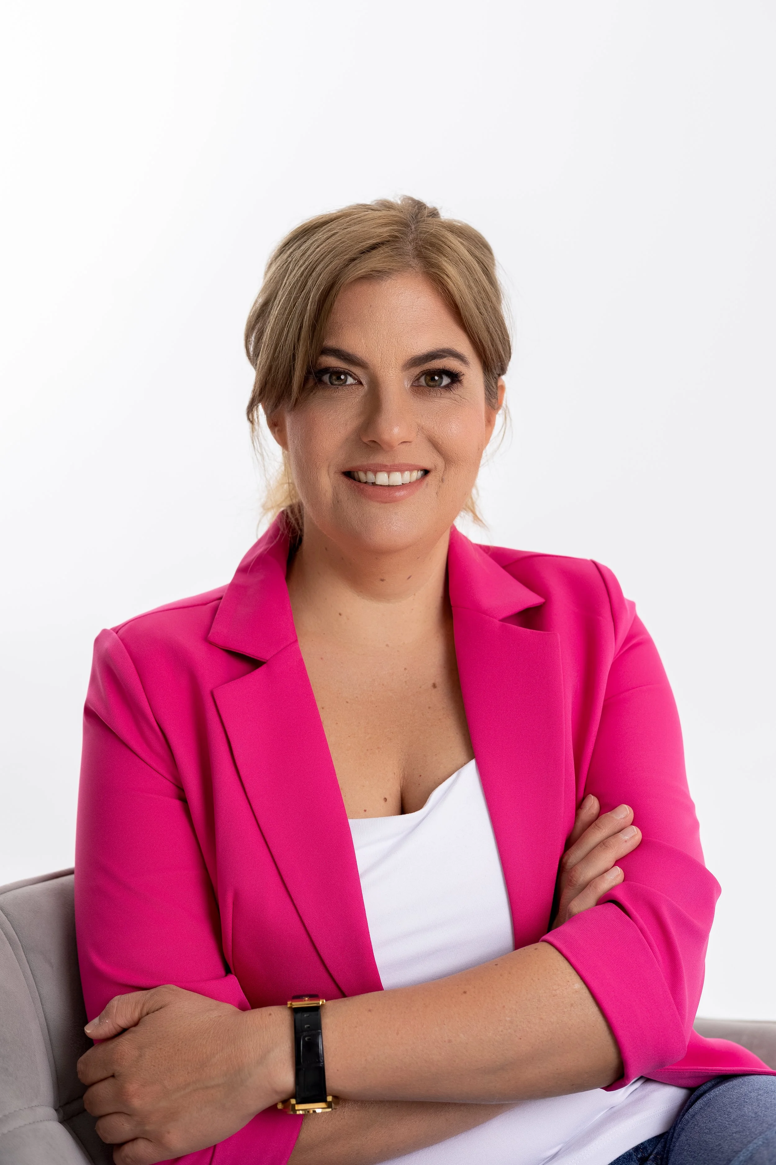Woman with blonde hair wearing a pink blazer and white top, sitting with arms crossed against a white background.