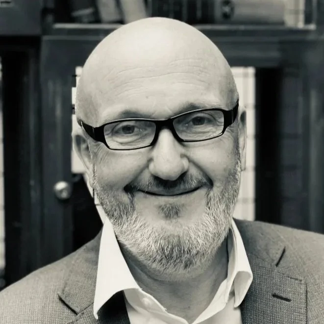 Close-up black-and-white photo of a smiling middle-aged man with glasses, a bald head, a beard, and wearing a suit jacket and white shirt.