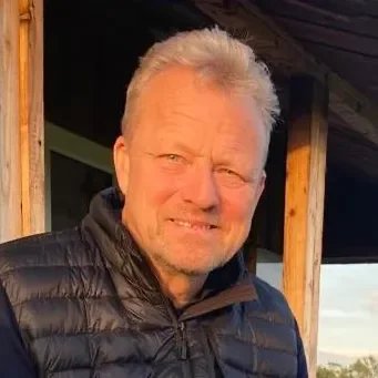 A smiling older man with gray hair, wearing a black puffer jacket, standing outdoors near a wooden structure.
