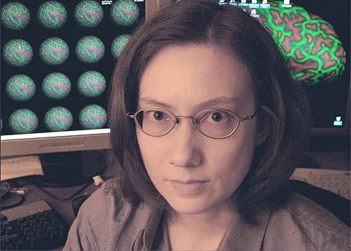 A woman with glasses in front of a dual-monitor computer setup displaying medical images of brains.