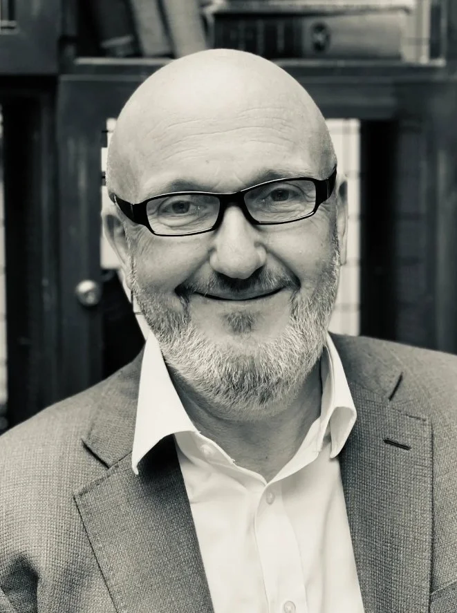 A smiling middle-aged man with glasses and a beard, wearing a suit jacket and white shirt, in a black and white photo.