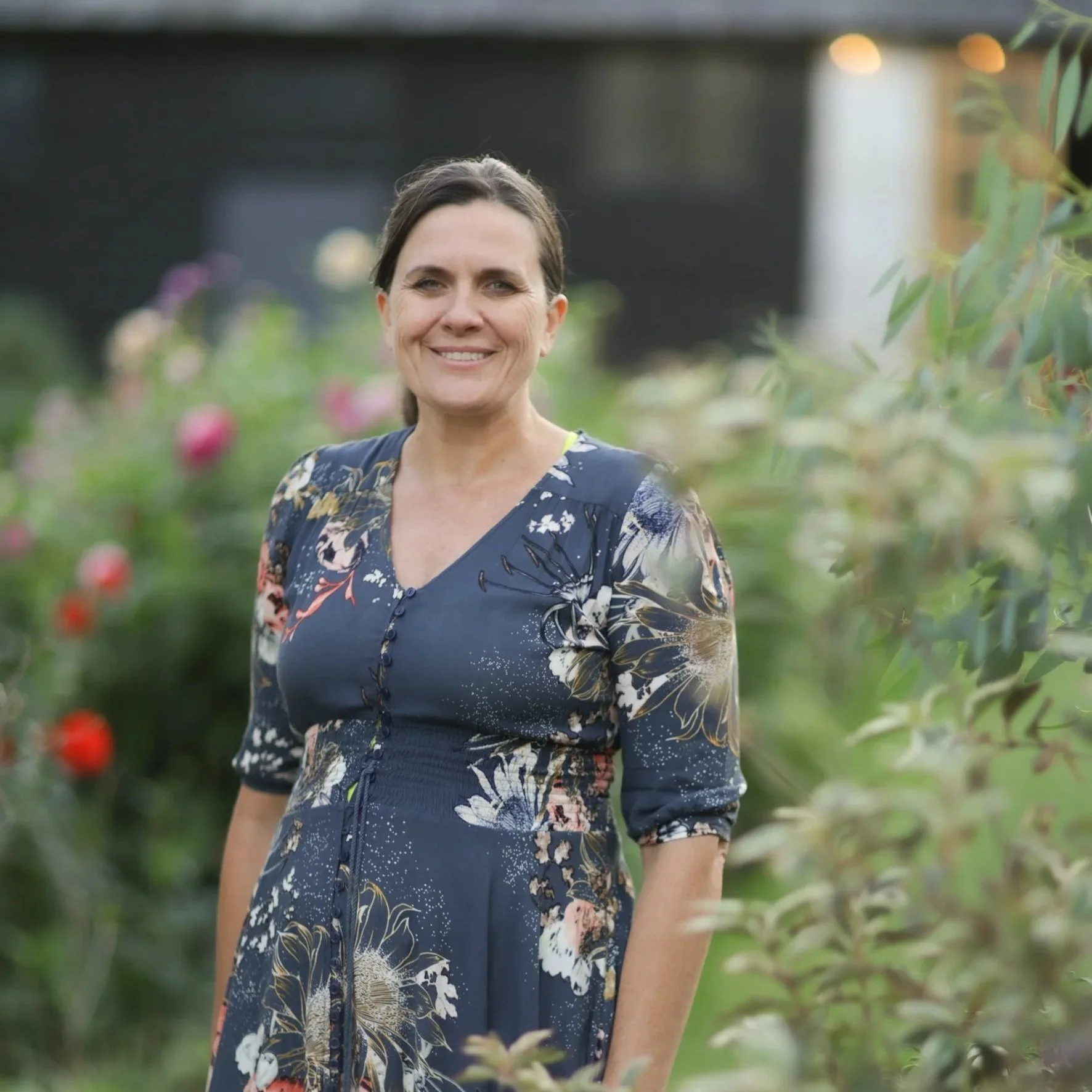 A woman in a floral dress smiling at the camera in a garden with blooming flowers.