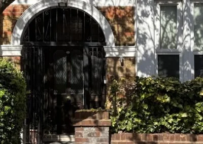 Black wrought iron gate with brick arch, surrounded by greenery and shadows from trees.