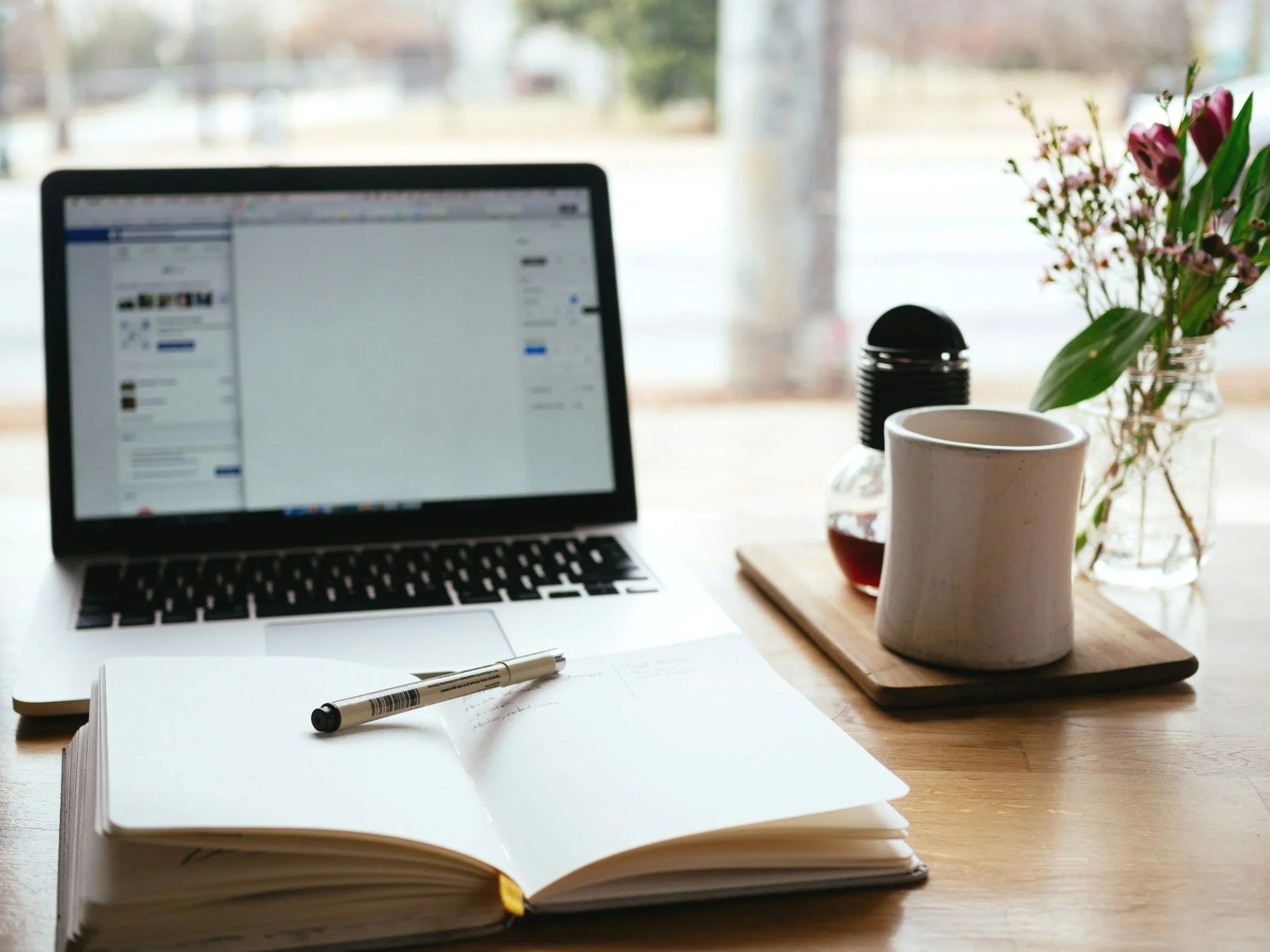 A workspace with a laptop displaying a social media page, an open notebook with a pen, a ceramic mug, a glass vase of flowers, a small black object, and a dining wooden tray. The workspace is near a window with a blurred view of trees and a park outside.