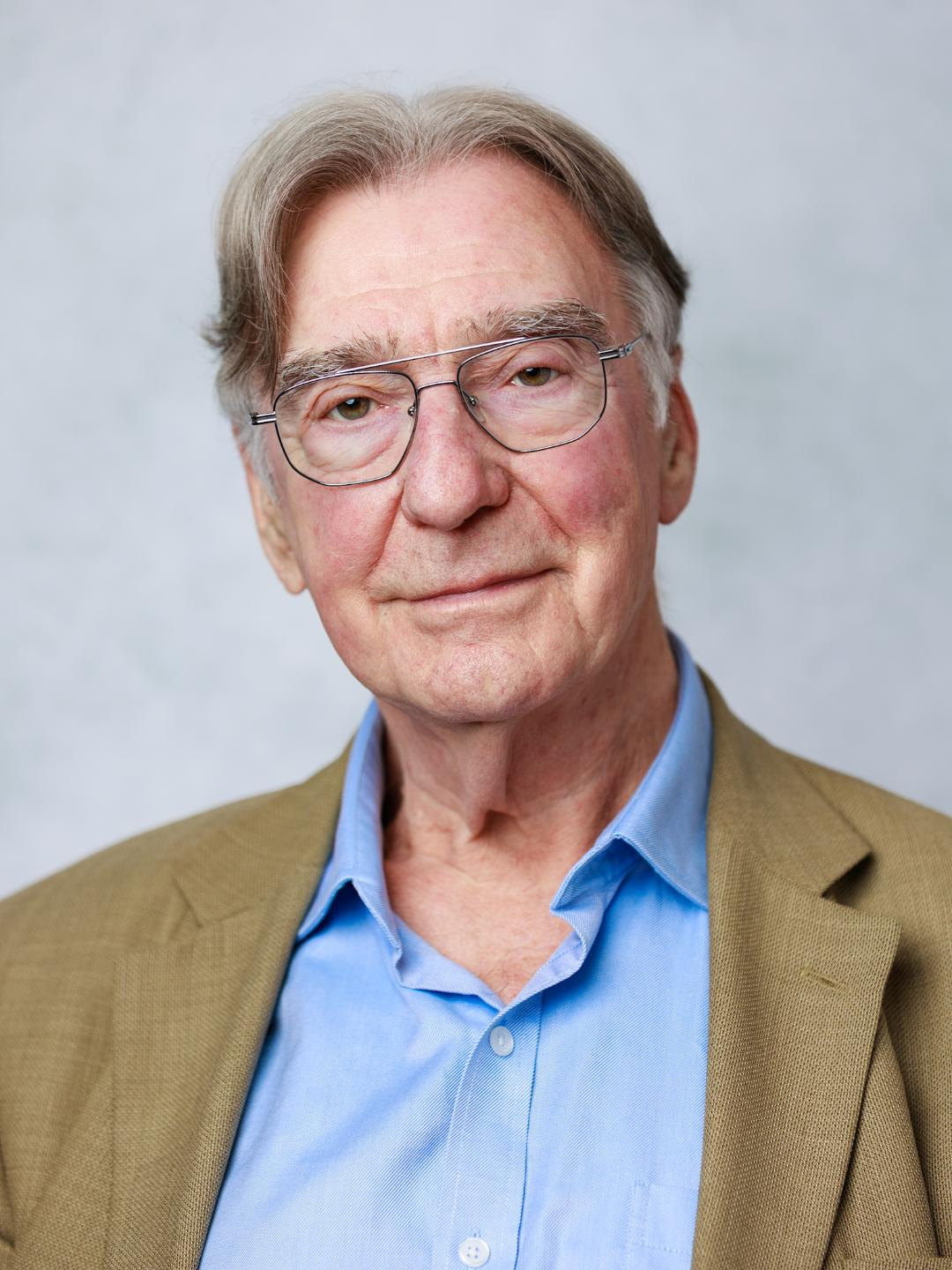 A middle-aged man with brown hair, glasses, and a light blue collared shirt smiling in front of a plain background.