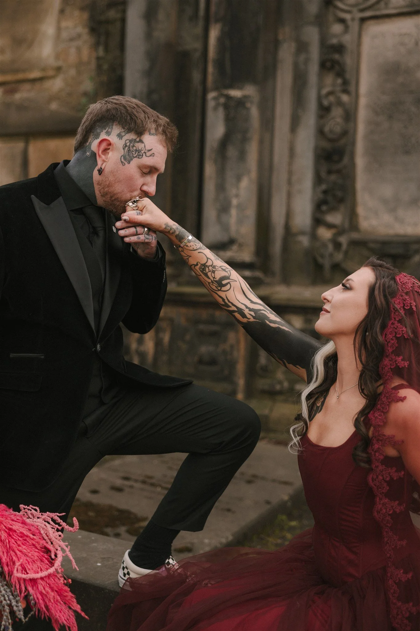 heavily tattooed groom dressed in black tie kissing the hand of his alternative bride, dressed in red, sitting on a greyfriar stone in edinburgh graveyard
