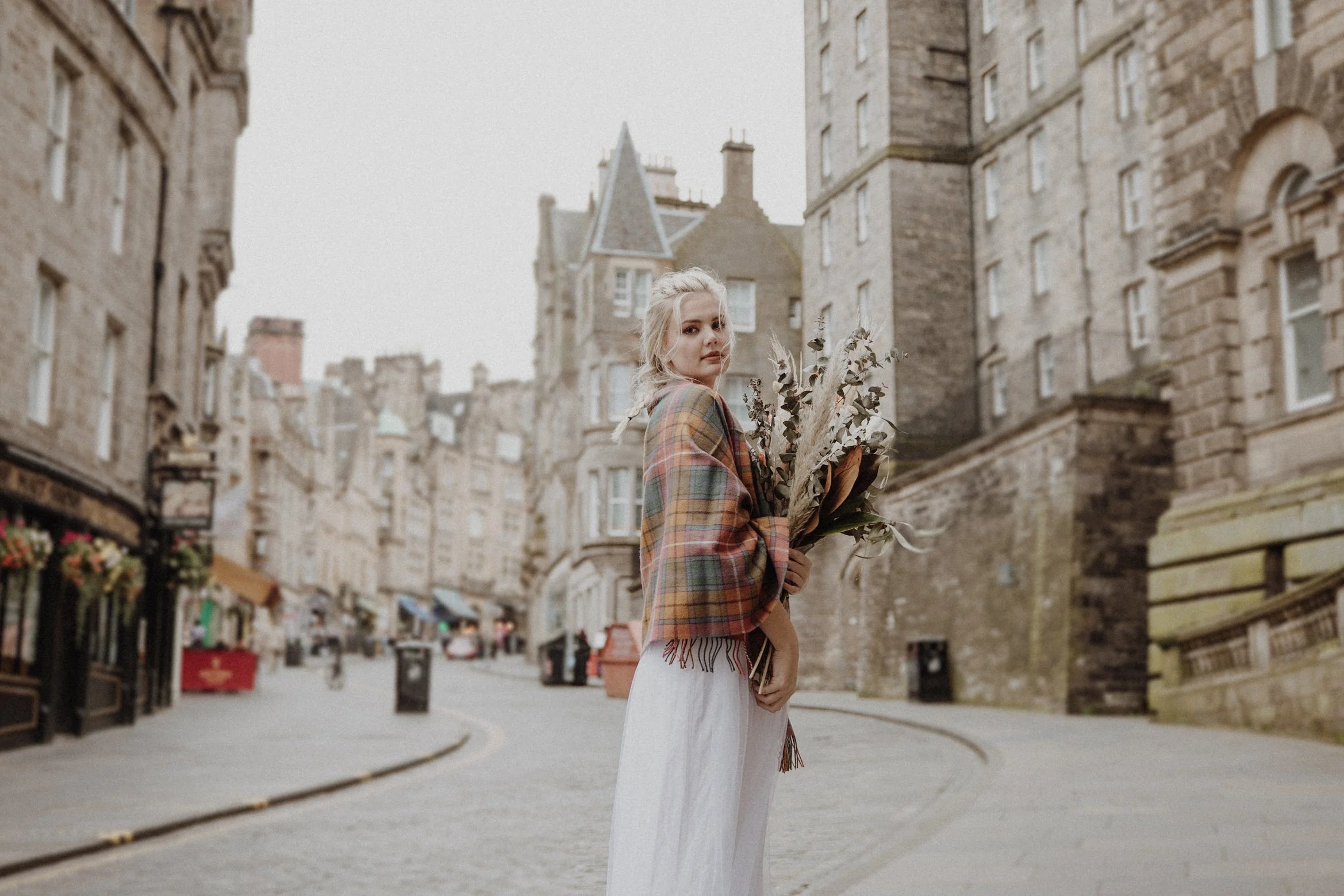  bridal photo portrait of blonde model walking in edinburgh old town Royal Mile with a bouquet of dried flowers in arm and edinburgh old town in the background 