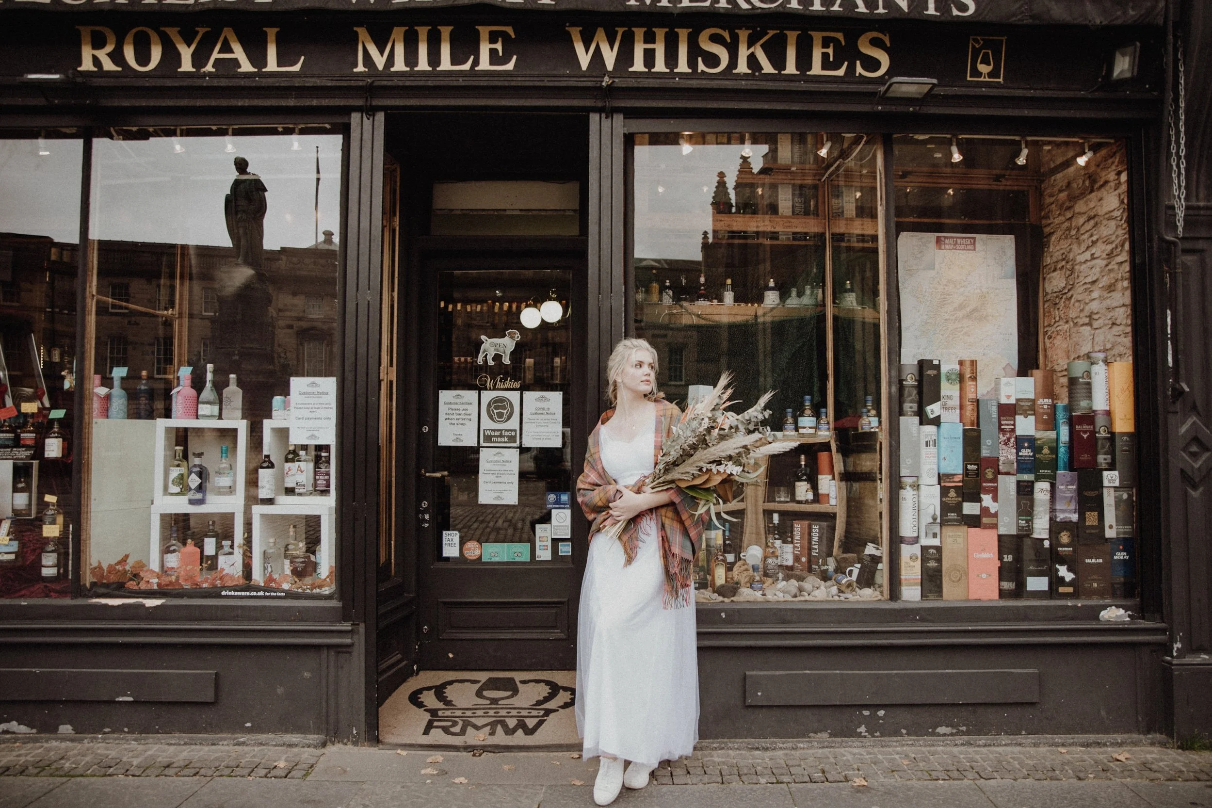  bridal photo portrait of blonde model walking in edinburgh old town Royal Mile with a light camel coat and a bouquet of dried flowers in arm 