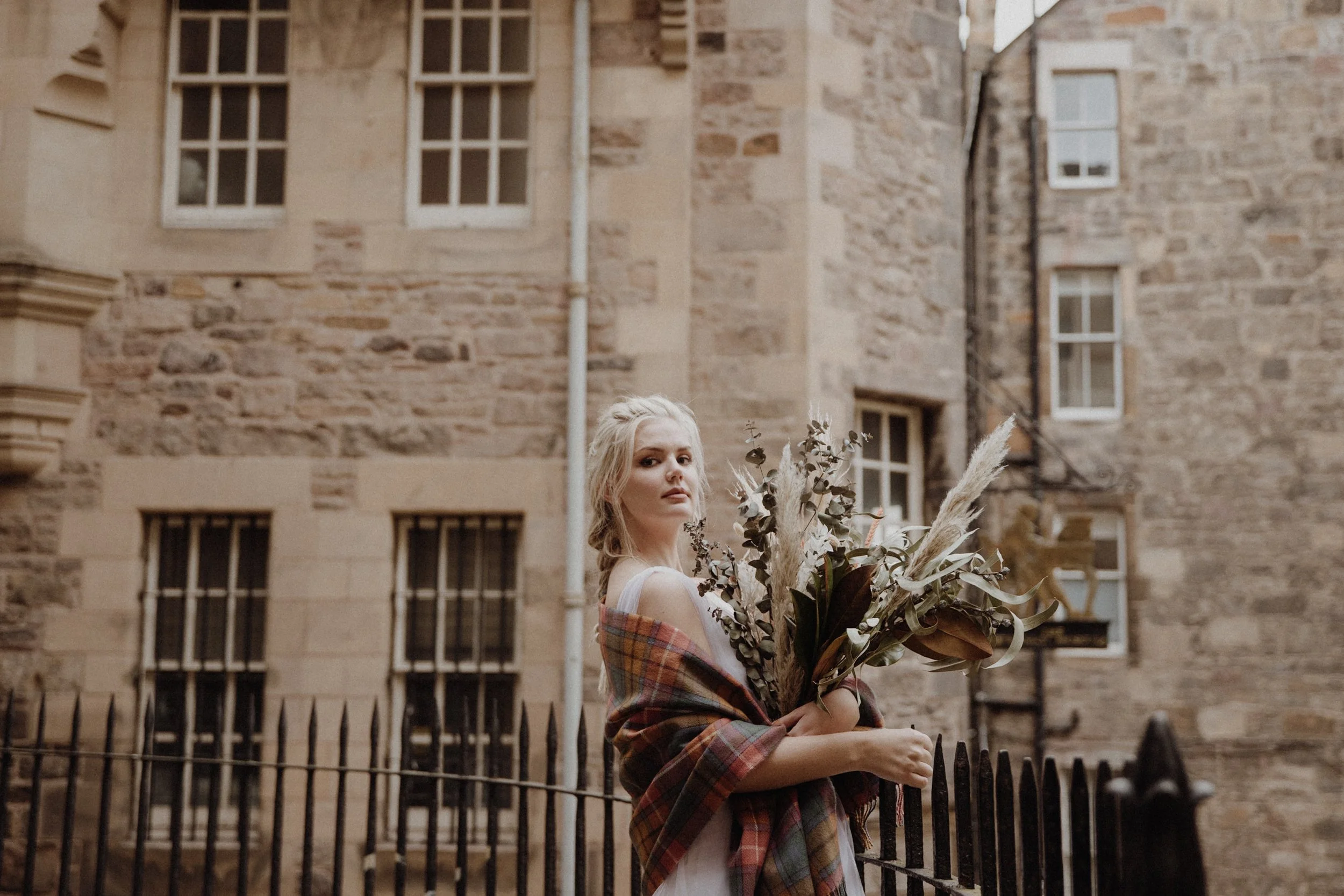  bridal photo portrait of blonde model walking in edinburgh old town Royal Mile with a light camel coat and a bouquet of dried flowers in arm 
