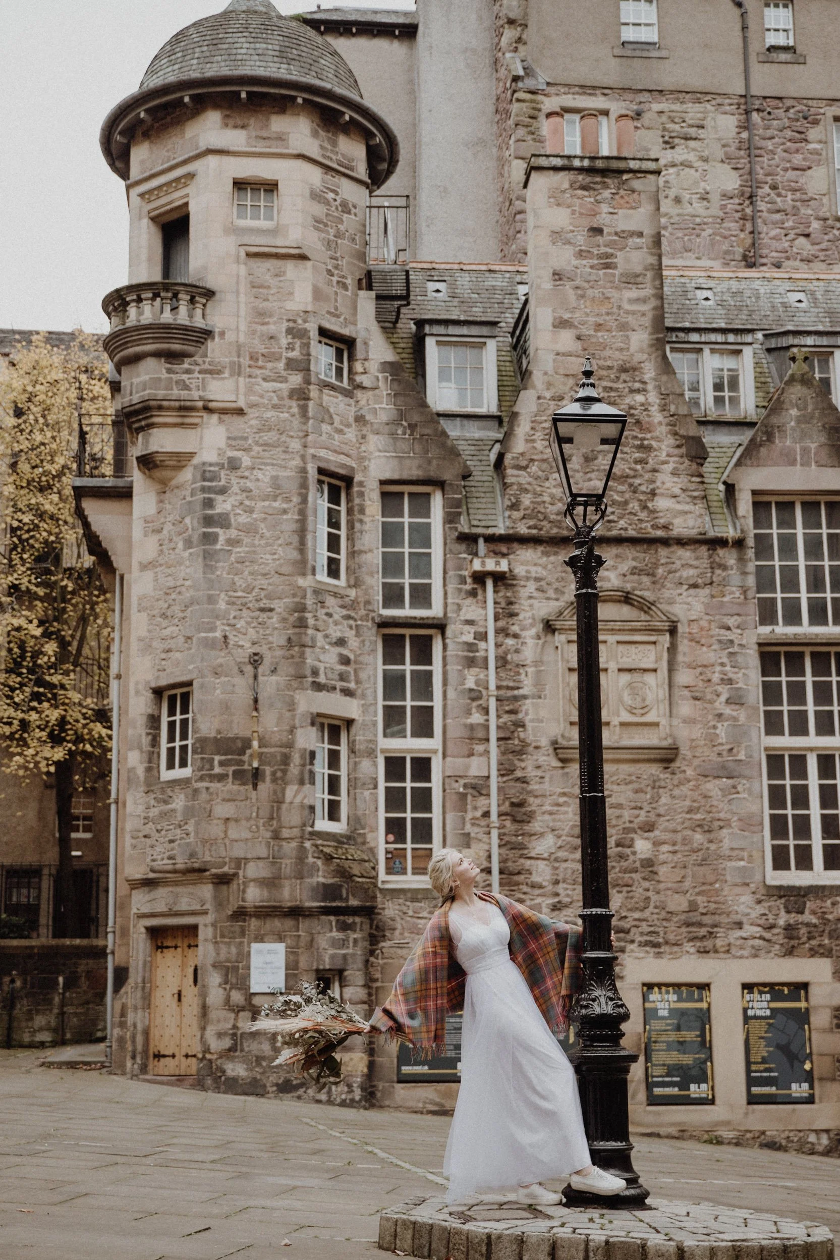  bridal photo portrait of blonde model walking in edinburgh old town Royal Mile with a light camel coat and a bouquet of dried flowers in arm 