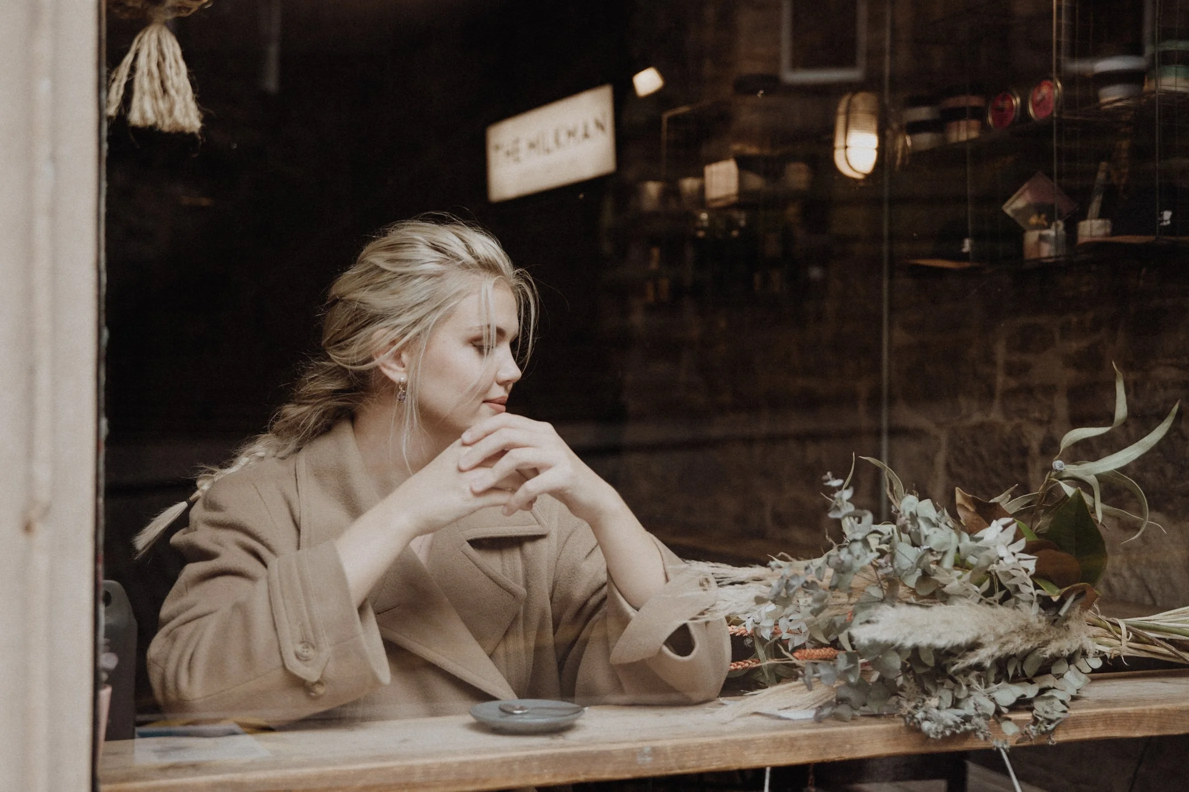  Bridal model with a white wedding gown and a camel coat sitting on the window of Milkman cafe in Edinburgh holding a coffee mug and looking outside, with a dried flower bouquet next to her 