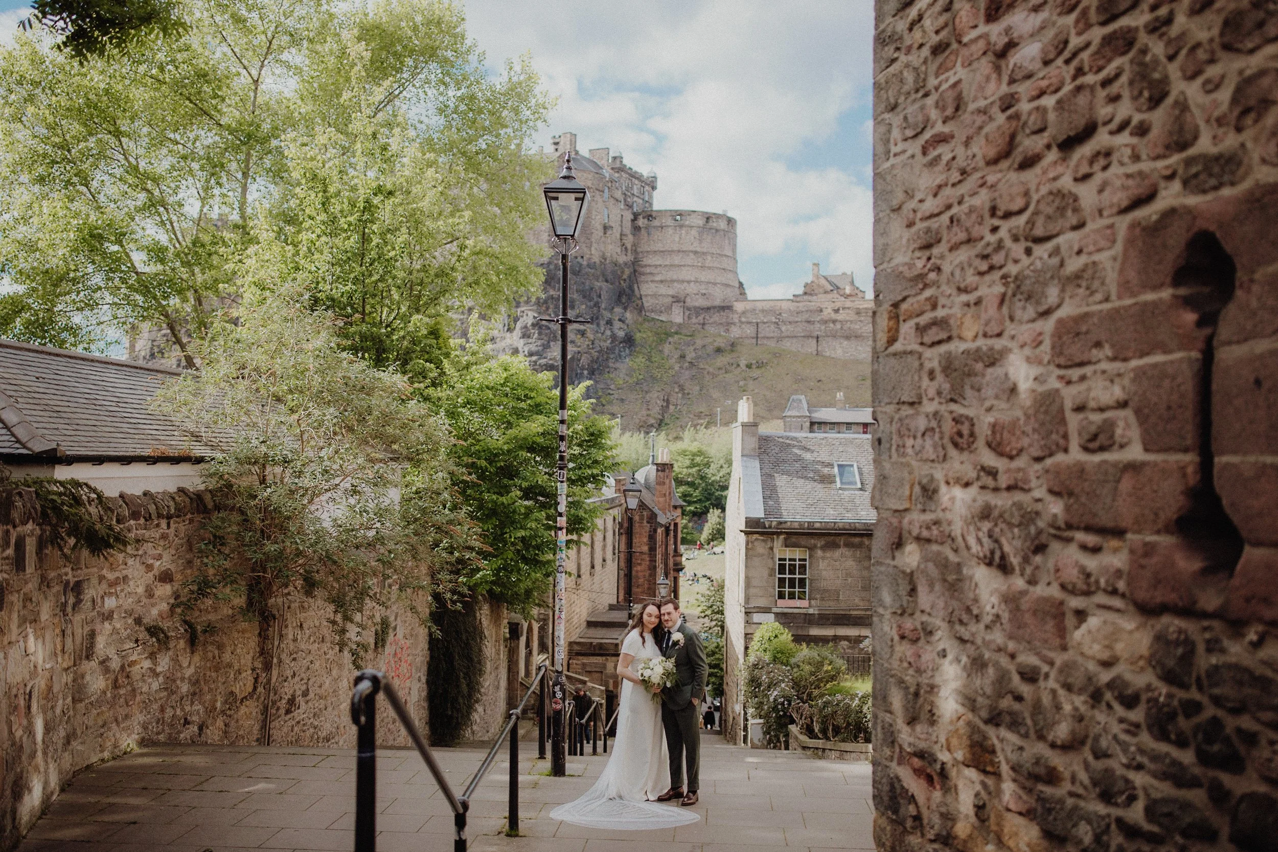 Bride and groom on the iconic fennel steps in Edinburgh with edinburgh castle in the background, stone walls around them and vintage lamp post next to them, standing hugging