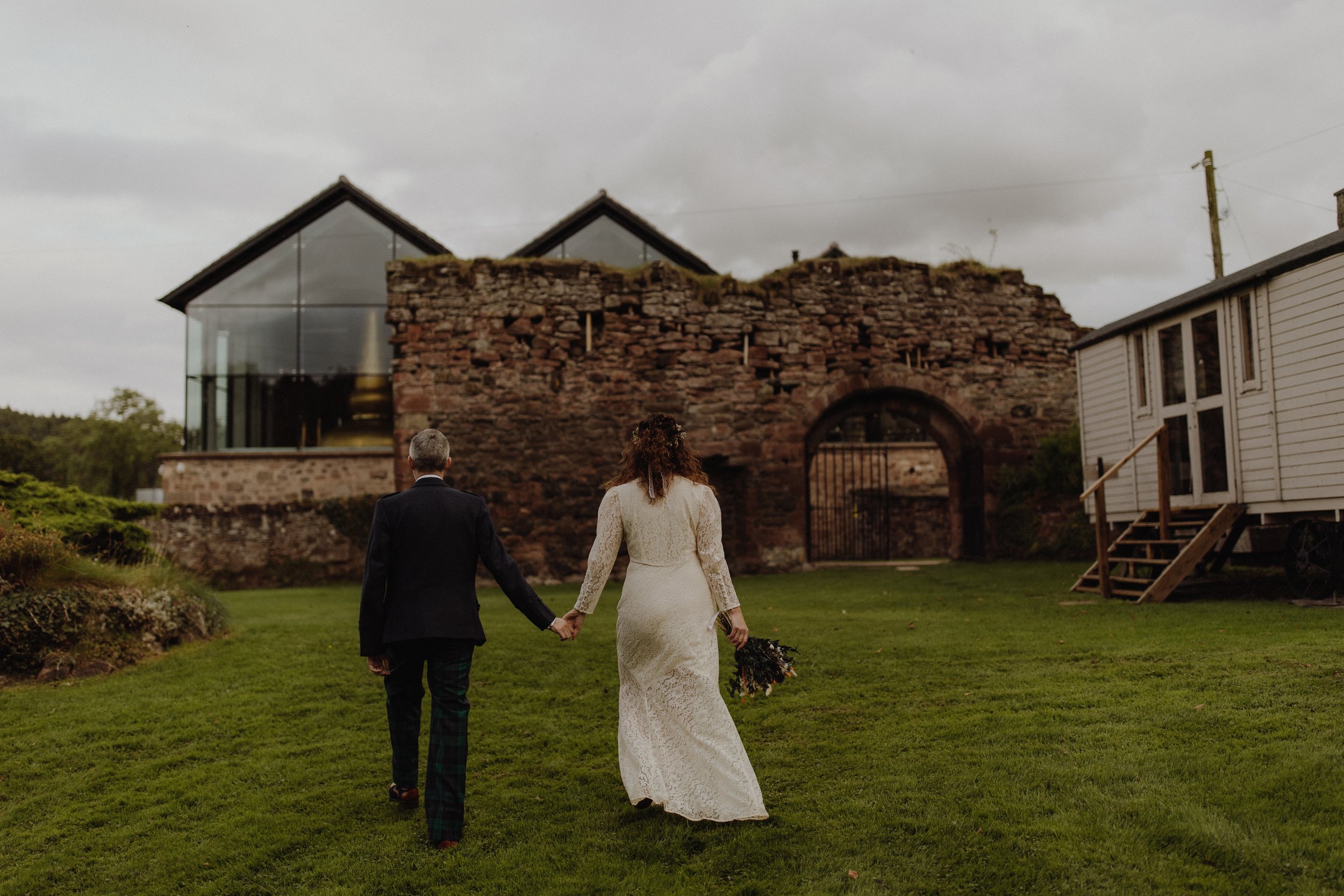 Wedding couple in wedding gowns taken from behind walking hand in hand in a an ancient welled garden towards lenders abbey distillery the distillery windows showing the copper pots