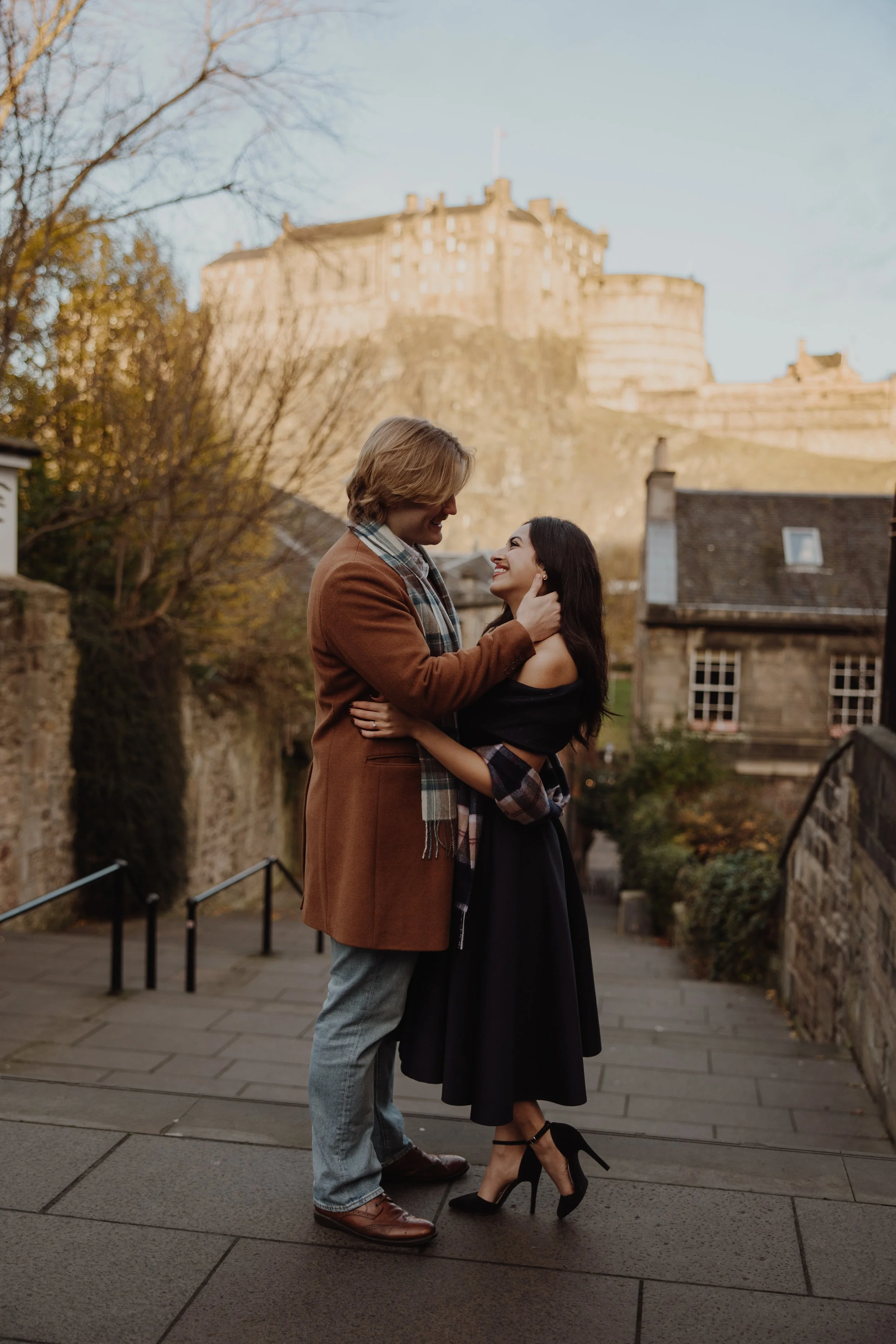 A couple embracing during a romantic engagement photoshoot at The Vennel in Edinburgh, with Edinburgh Castle glowing in the background above Grassmarket, framed by historic stone buildings and soft autumn light.
