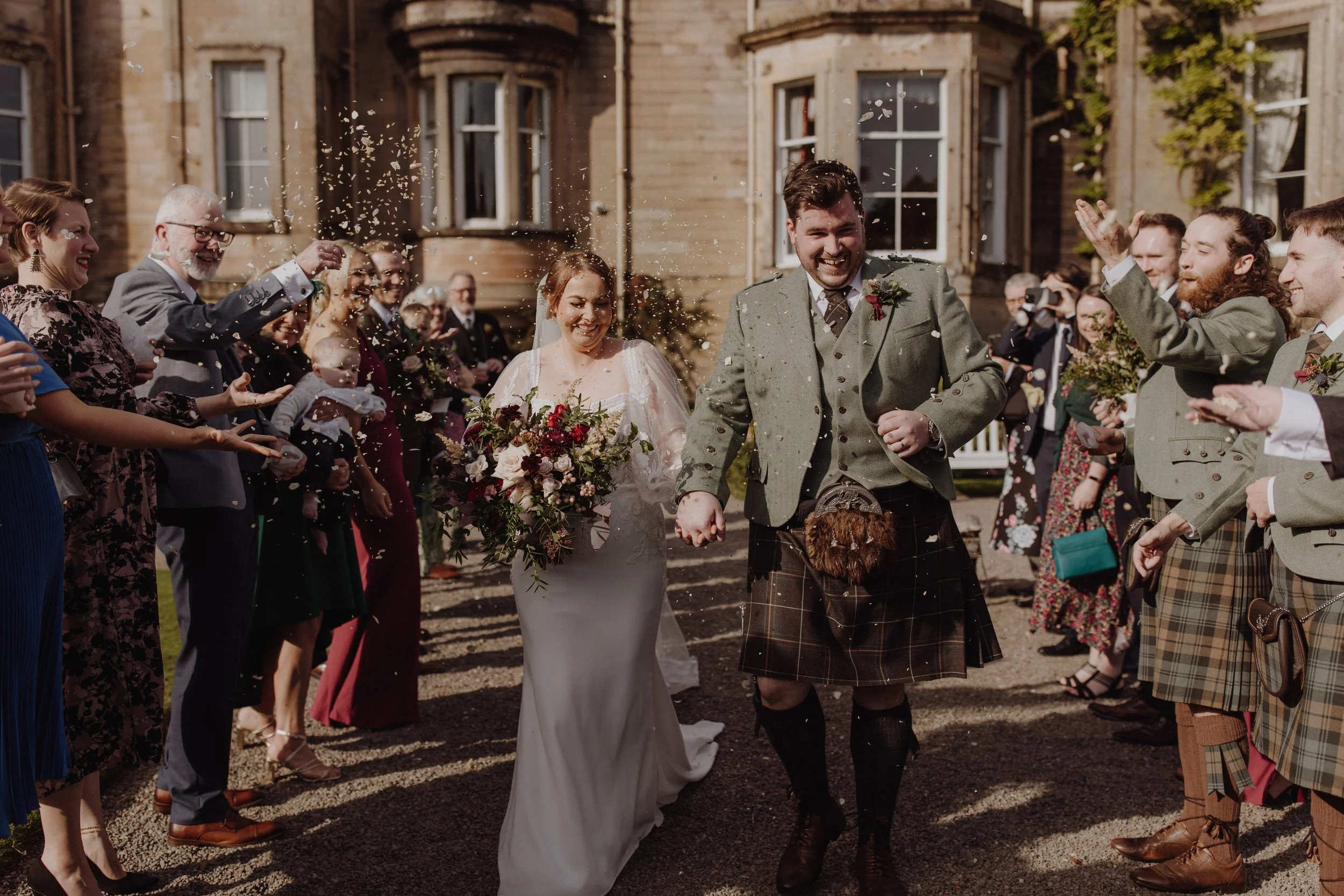 Bride and groom in traditional Scottish tartan kilt suit walking between their guests forming a tunnel while throwing dried petal confetti in front of auchen castle