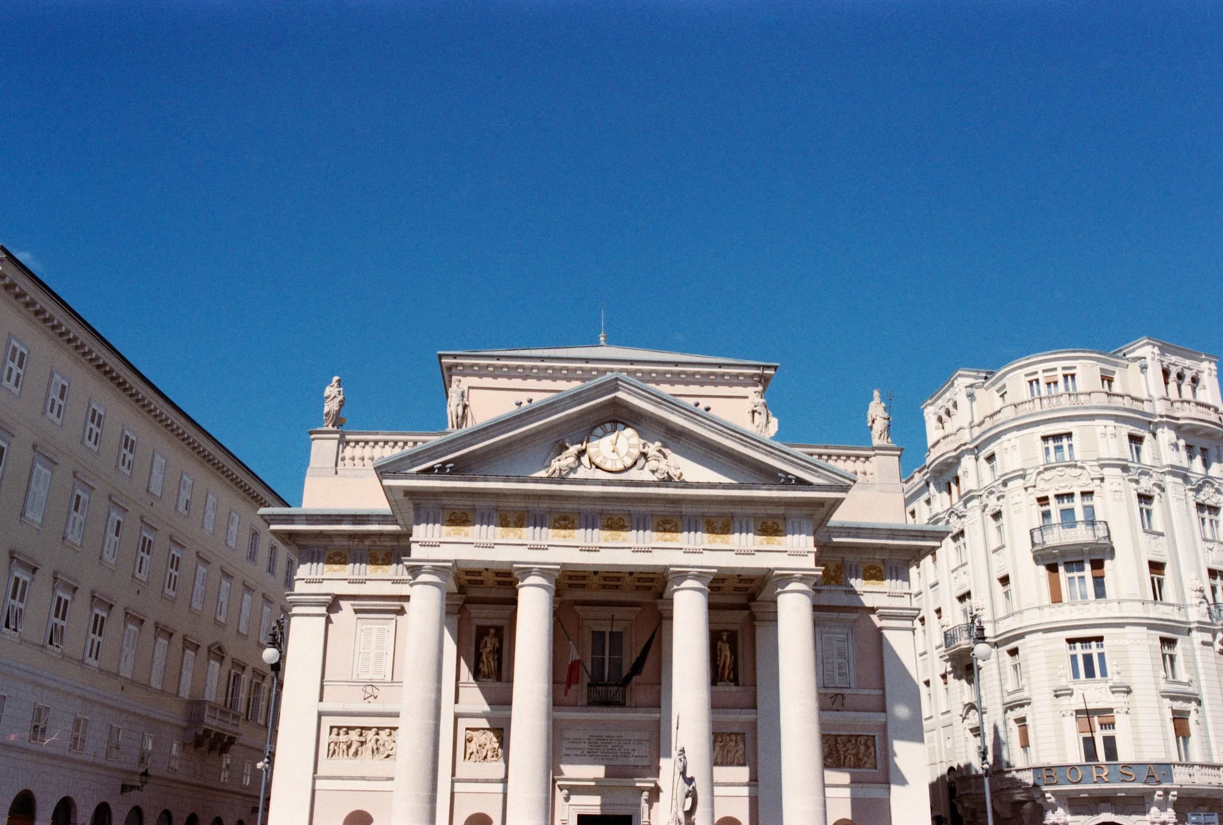 Piazza della Borsa in Trieste showing A historic neoclassical building with columns and statues on the roof, situated between modern multi-story buildings, against a clear blue sky, taken on 35mm film