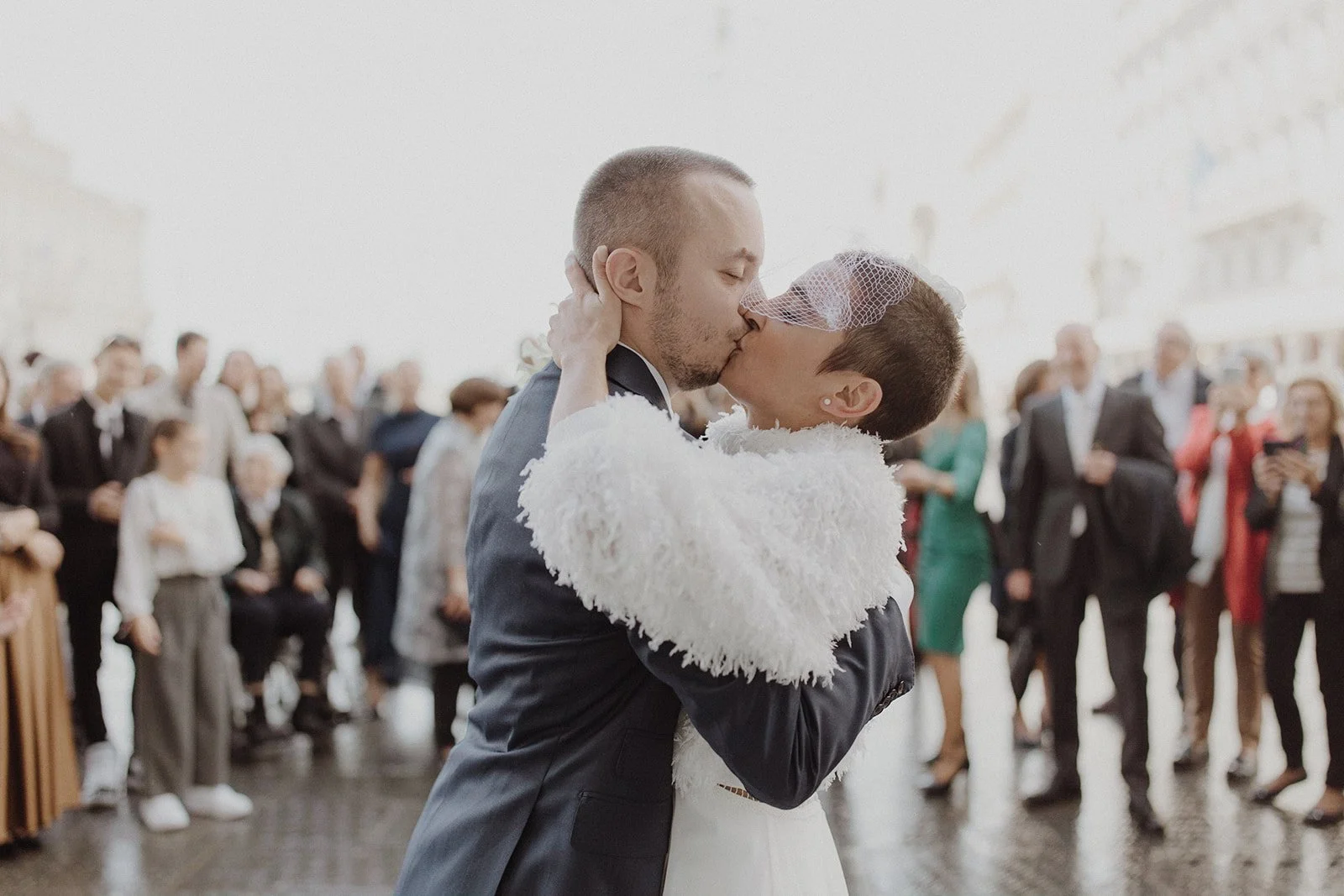 couple with alternative shaved head bride kissing in piazza unita di Italia in trieste with all their guests blurred in the background