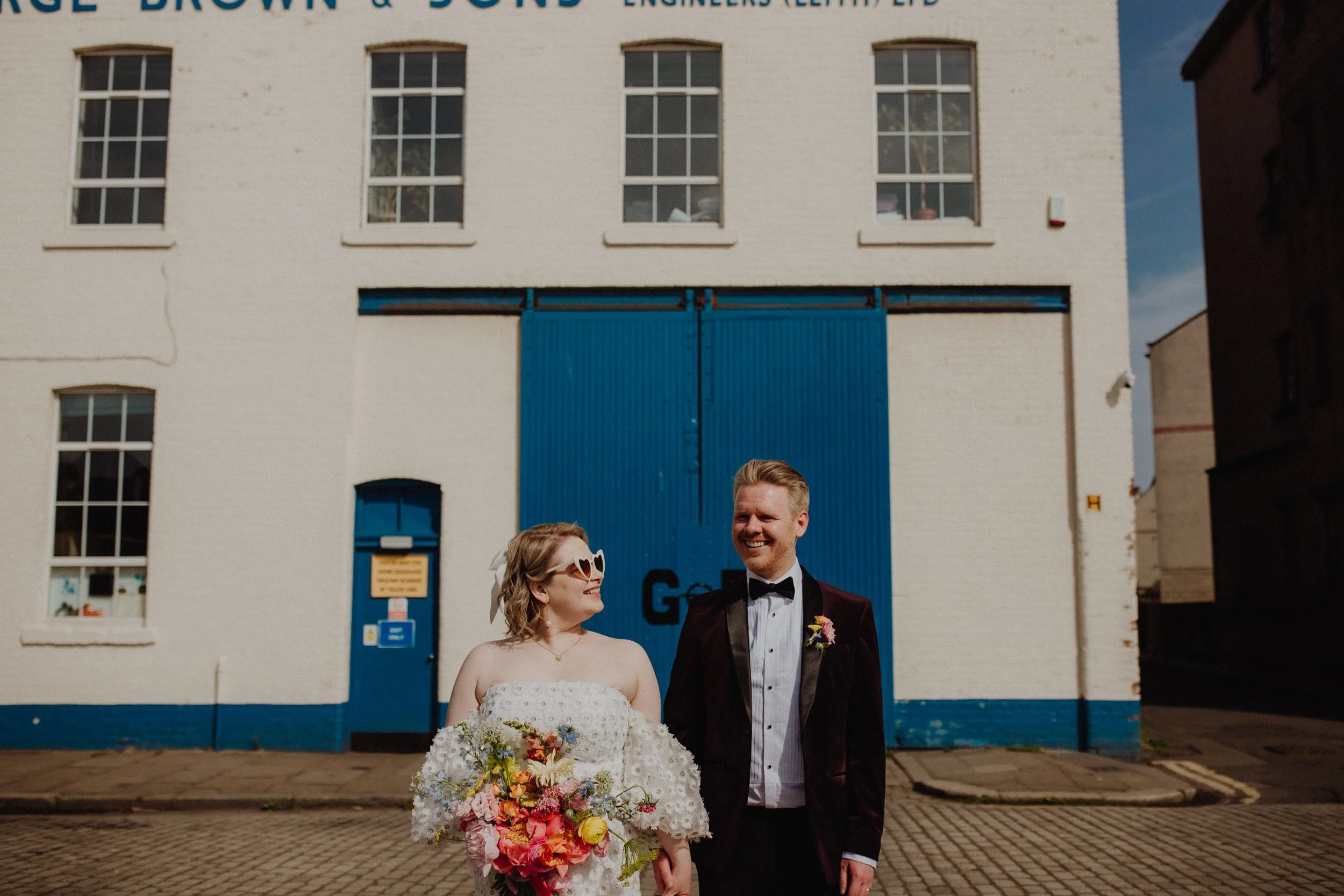 Alternative wedding couple with Fruit salad flowers bouquet bride in a daisy dress and sunglasses holding hands with smiling groom in front of Browns in Leith industrial blue doors