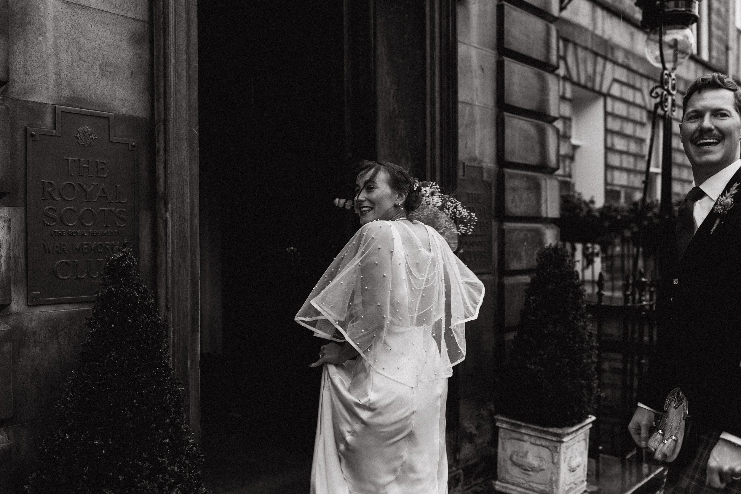 A bride in a wedding dress and veil smiles as she walks out of a building while a man in a suit, holding a small decorative object, stands nearby. The building has a sign reading "The Royal Scots" and a stone wall with planters at the entrance.