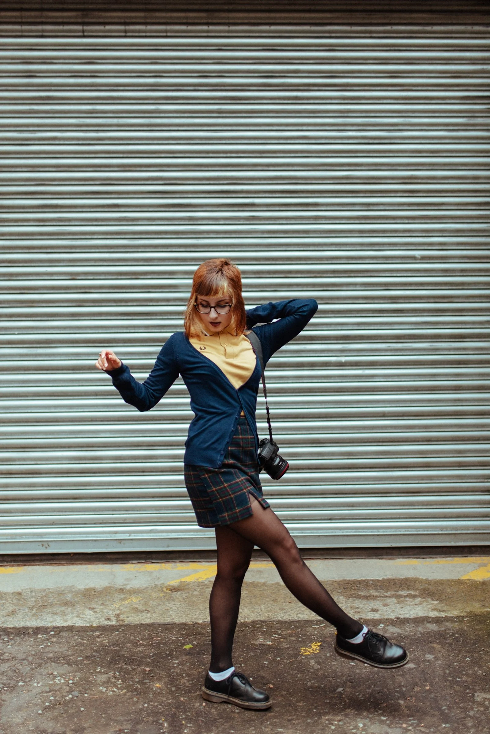 A young woman with glasses, wearing a navy cardigan, yellow top, plaid skirt, black tights, and black shoes with white socks, posing in front of a closed metal shutter with a camera hanging around her neck.