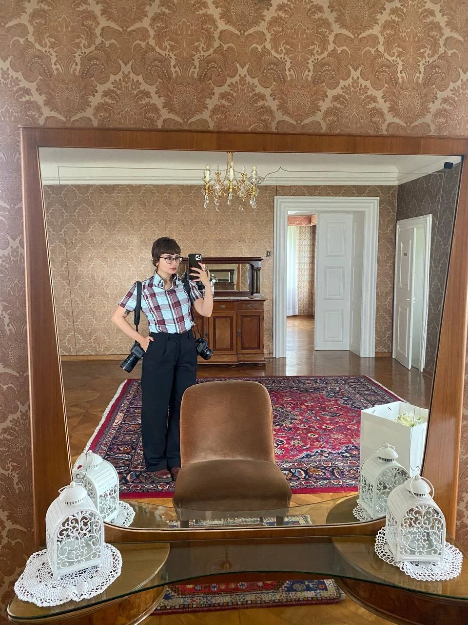 A woman taking a selfie in a large mirror in a vintage-style room. She has short hair, glasses, and is dressed in a plaid shirt and black pants. She has cameras hanging from her neck. The room has patterned wallpaper, a chandelier, a wooden sideboard, and a red patterned rug on the parquet floor. The mirror is decorated with two white lanterns and a glass table with white doilies.