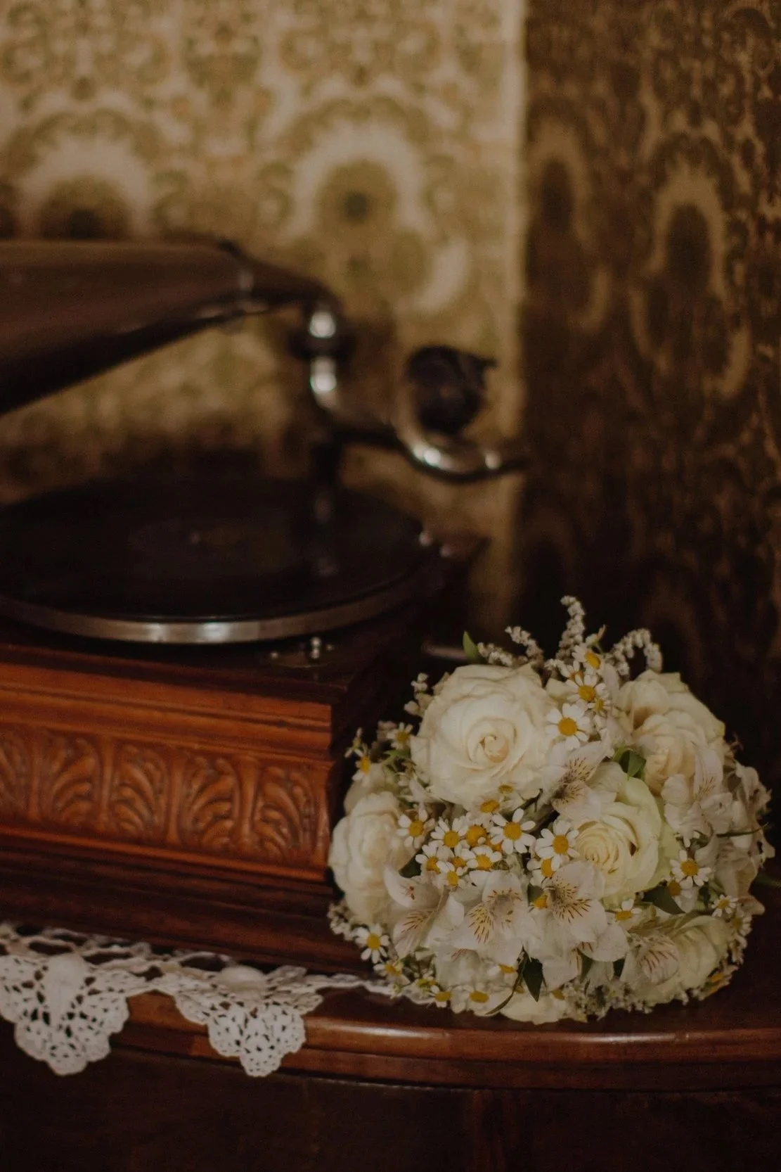 A vintage record player on a wooden table next to a bouquet of white roses and small daisies, with a decorative vintage lace cloth underneath.